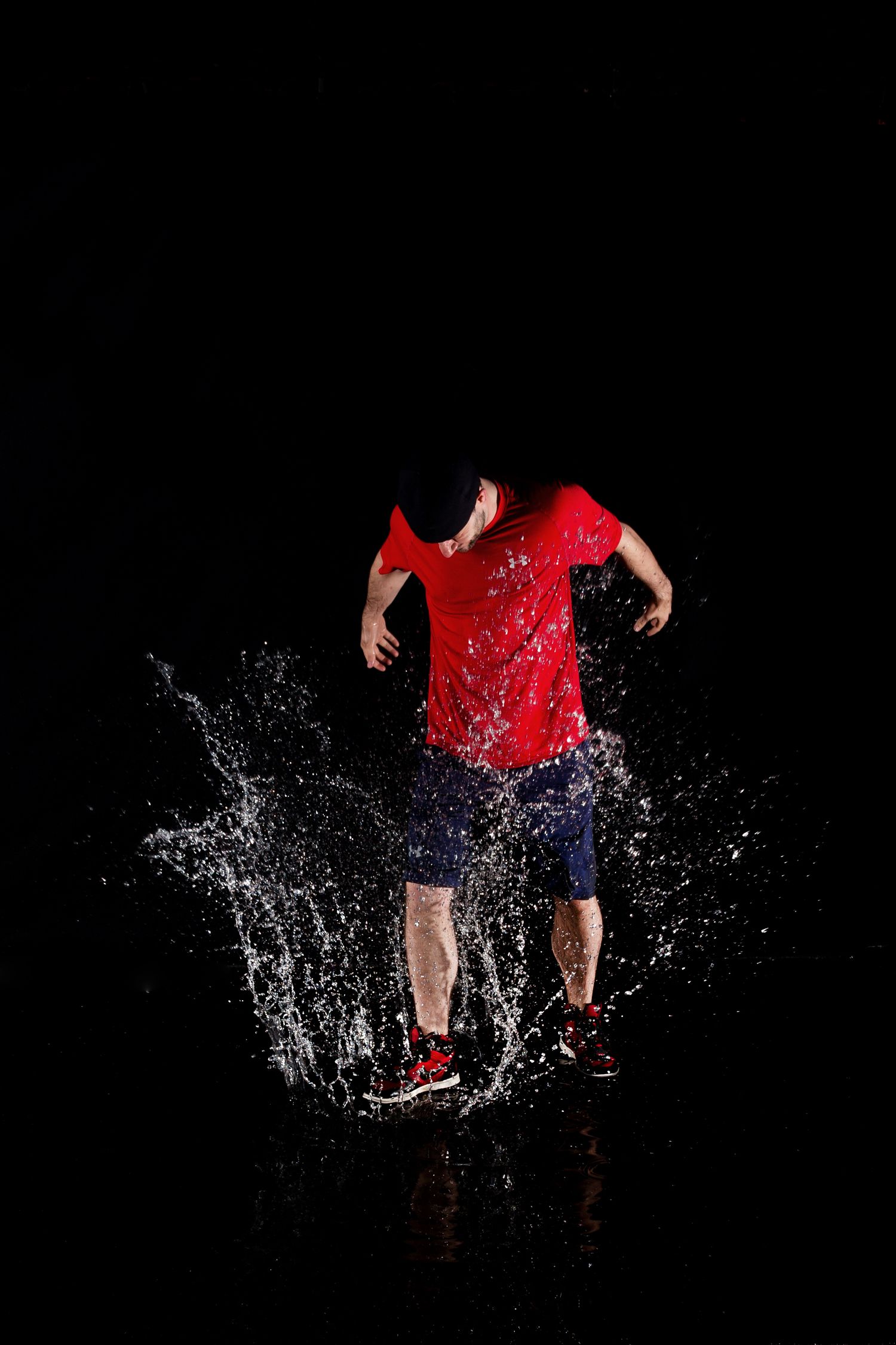 Person in red shirt and shorts jumping through water splashes against black background creating dramatic effect.