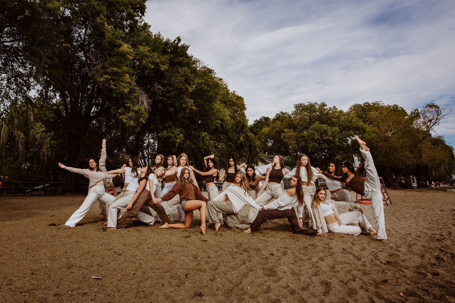 Group in all white clothing poses energetically together on sandy beach area with trees in background.