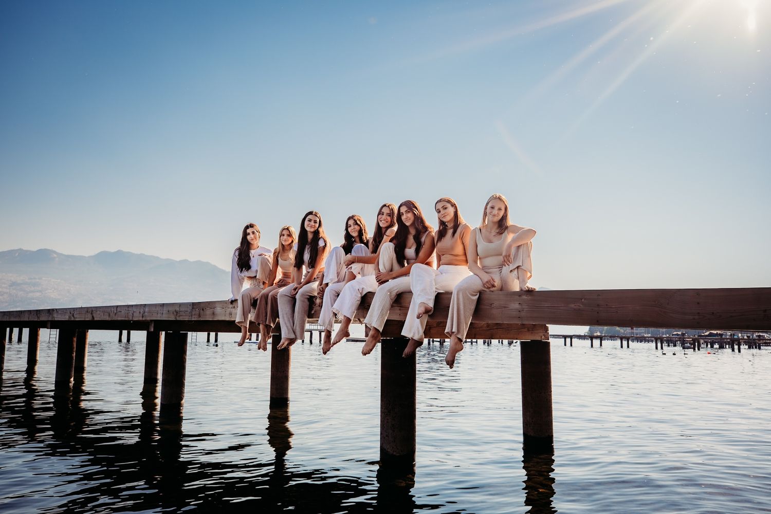 A group sits together on a wooden pier with dangling feet over calm lake water as sunlight streams through blue sky.