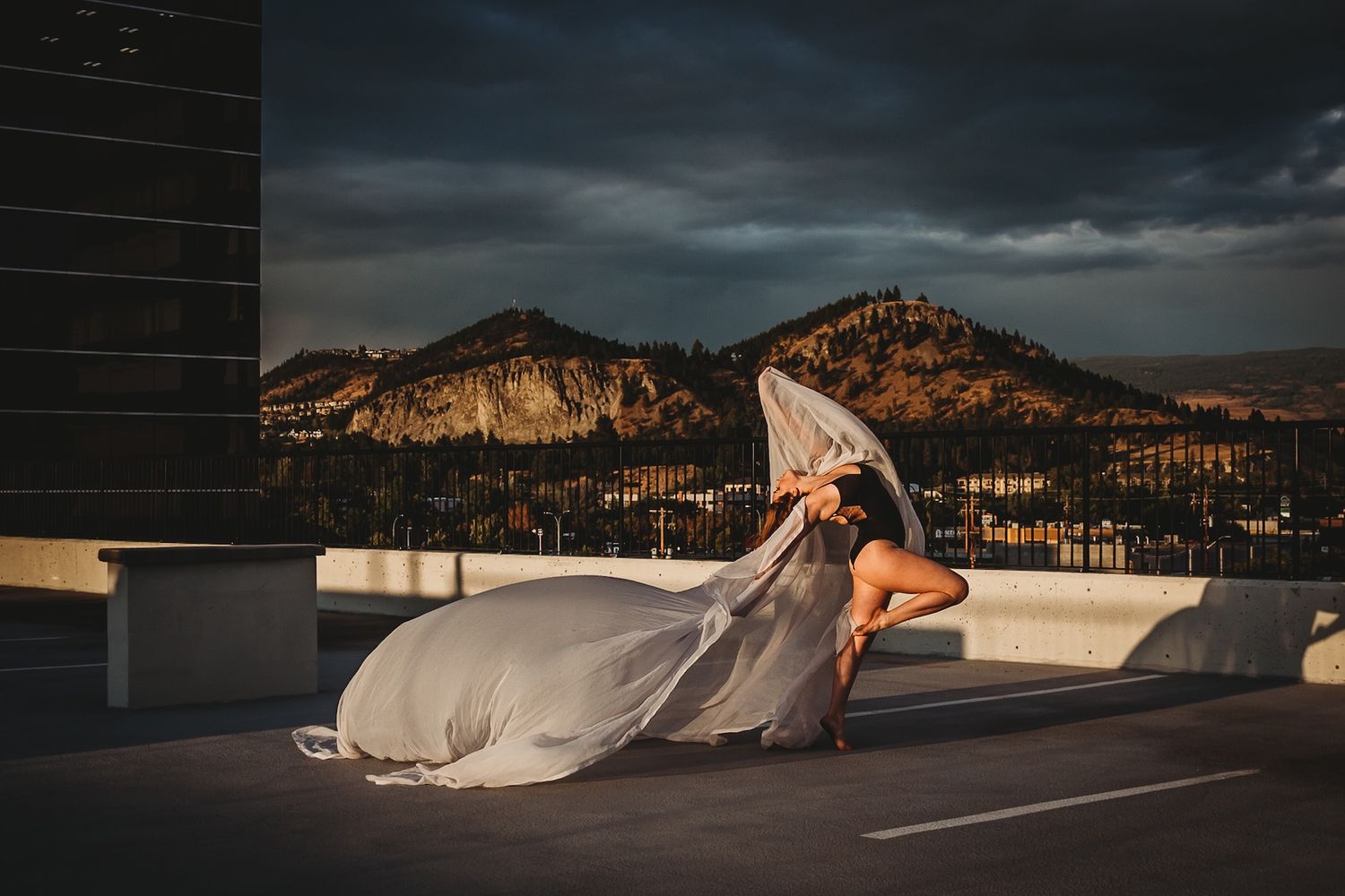 A flowing wedding dress billows dramatically on a rooftop parking deck with mountain peaks and dramatic clouds at sunset.