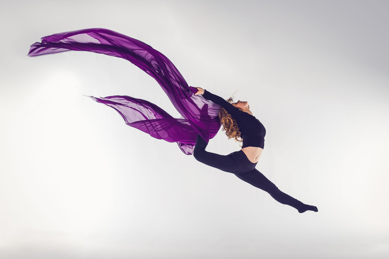 Dancer in black leotard leaps gracefully with flowing purple fabric trailing behind against light background.