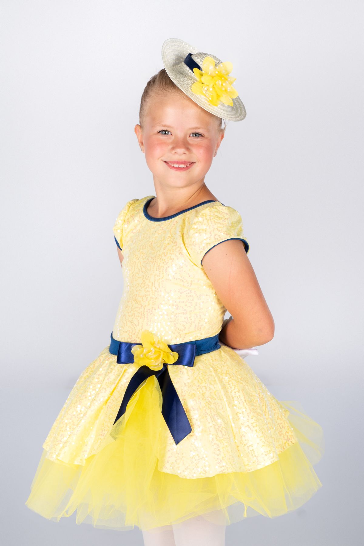 Young dancer wearing a yellow tulle dress with navy blue sash and small decorative hat performs in studio setting.