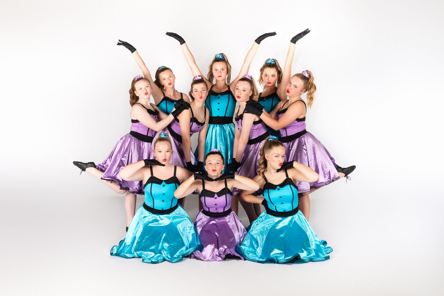 Dance group strikes dynamic pose wearing coordinated turquoise and purple dresses against white background.