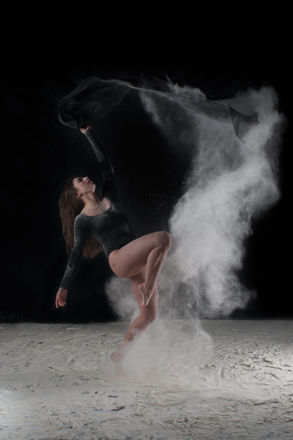 Athletic dancer leaps through swirling white powder creating ethereal effect against dark studio backdrop.