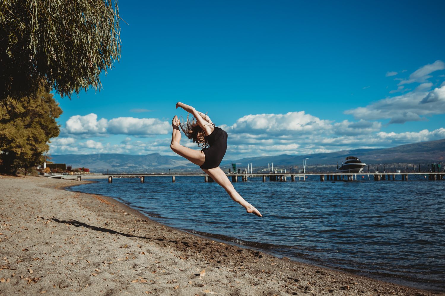 A dancer performs a graceful leap on a beach shoreline with a dock and mountains visible in the background.