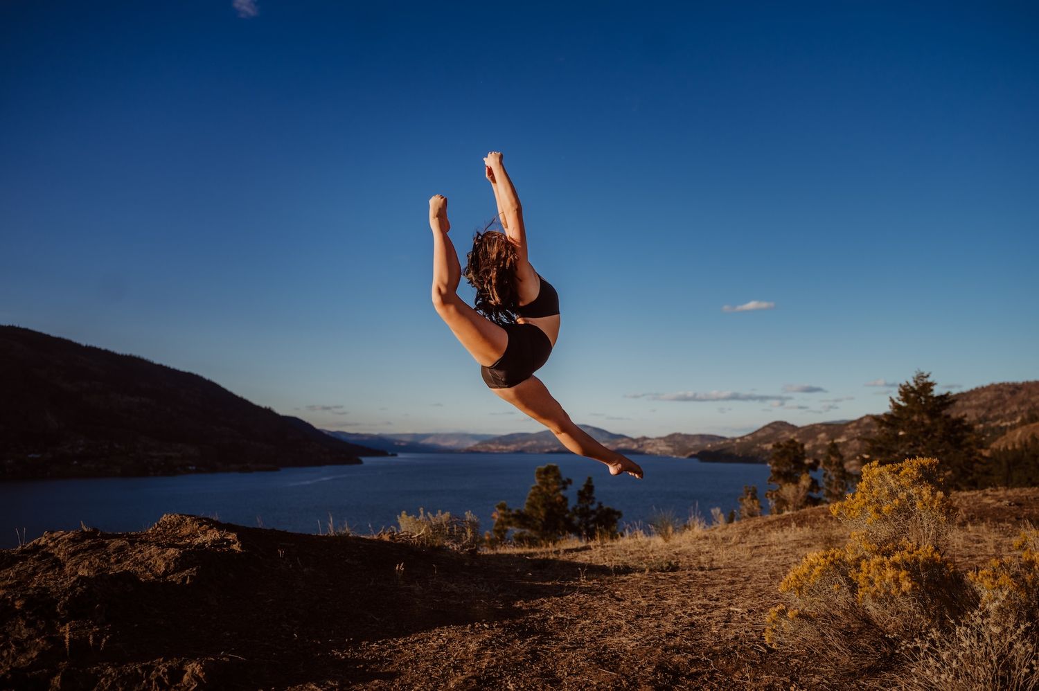 Graceful dancer performs a dramatic leap against a scenic mountain lake backdrop during golden sunset.