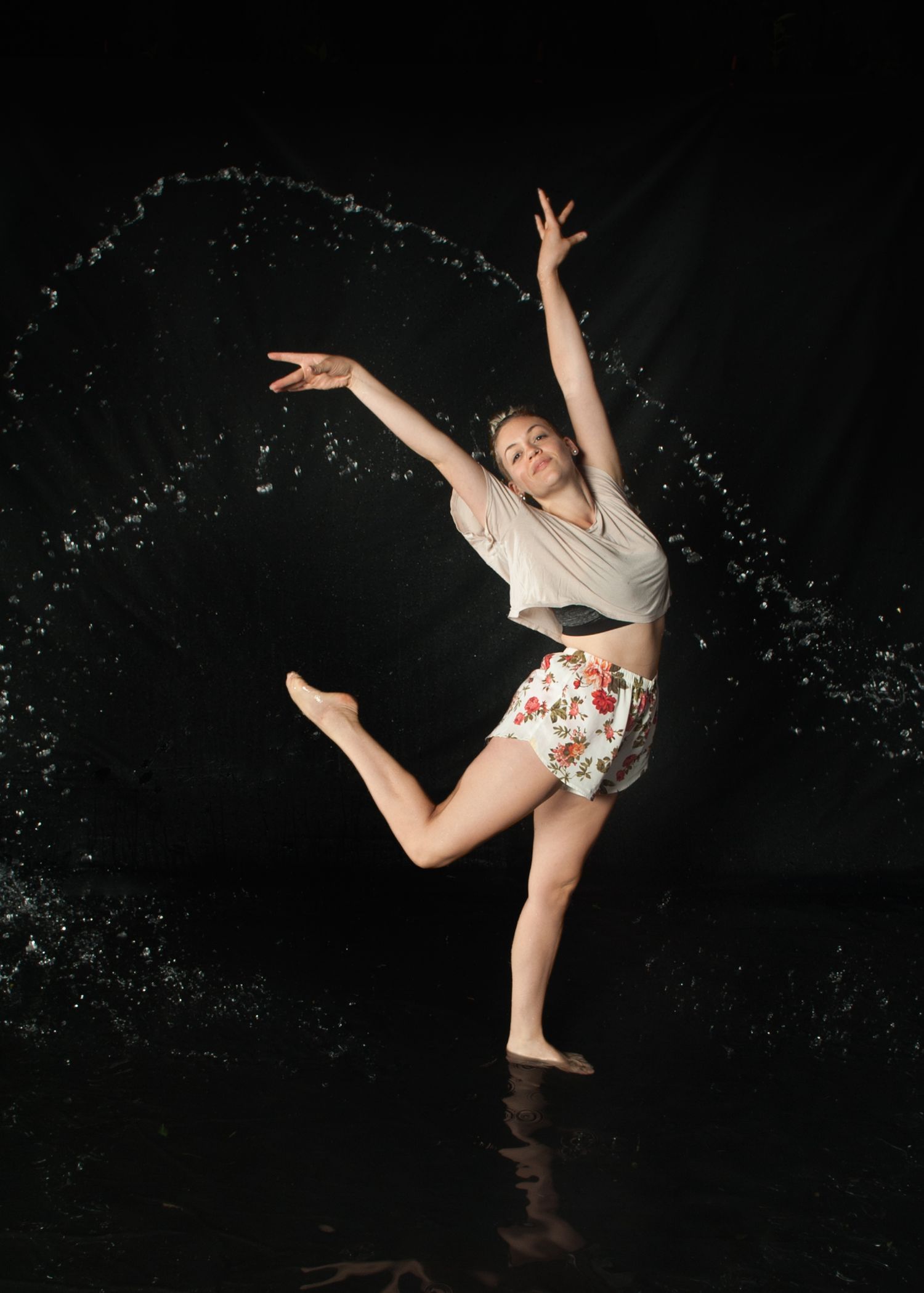 Contemporary dancer in floral shorts and white top makes graceful leap with water splashing against dark background.