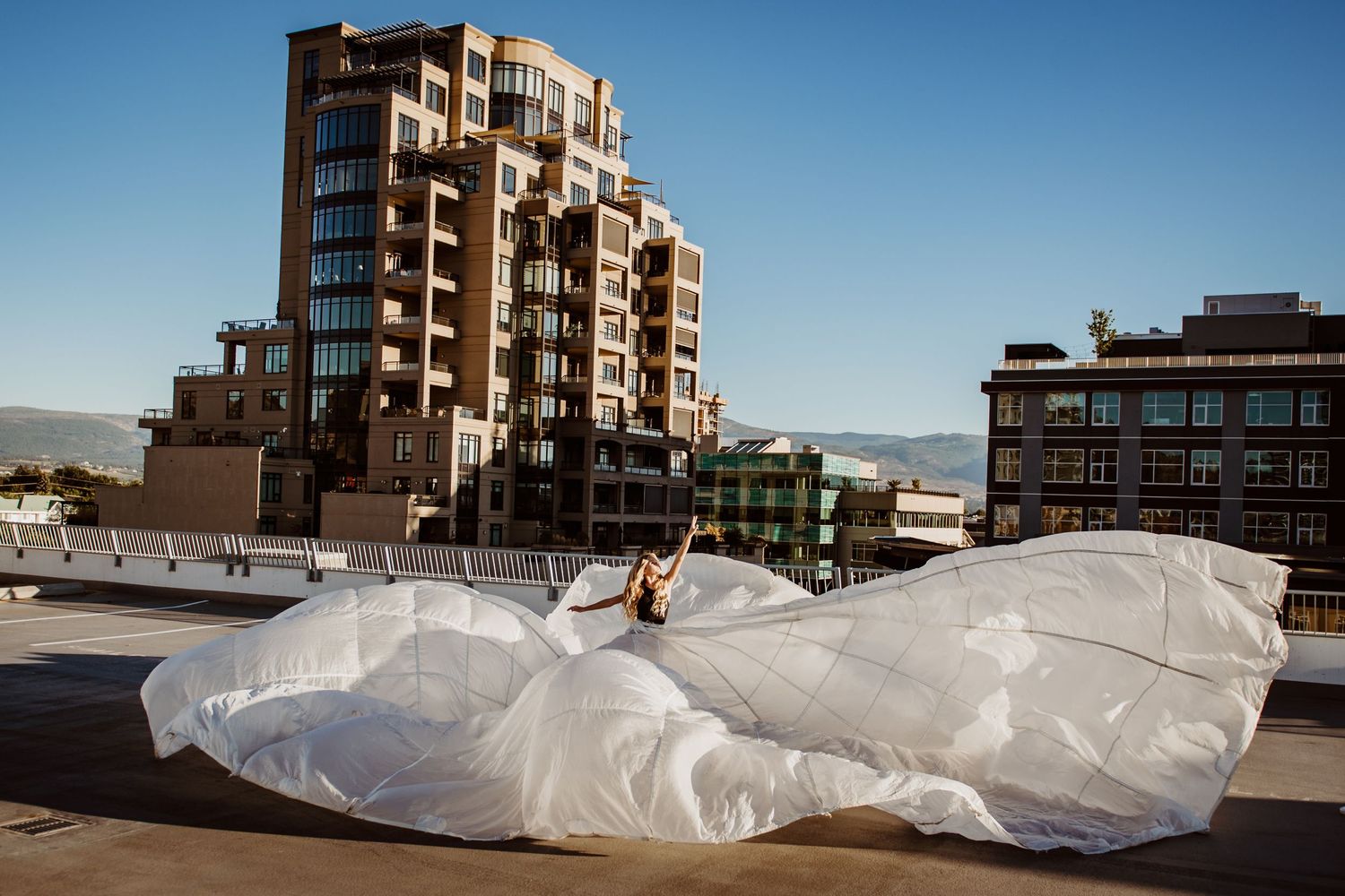 Someone in a flowing white wedding dress lying on a rooftop with urban buildings and mountains in the background.