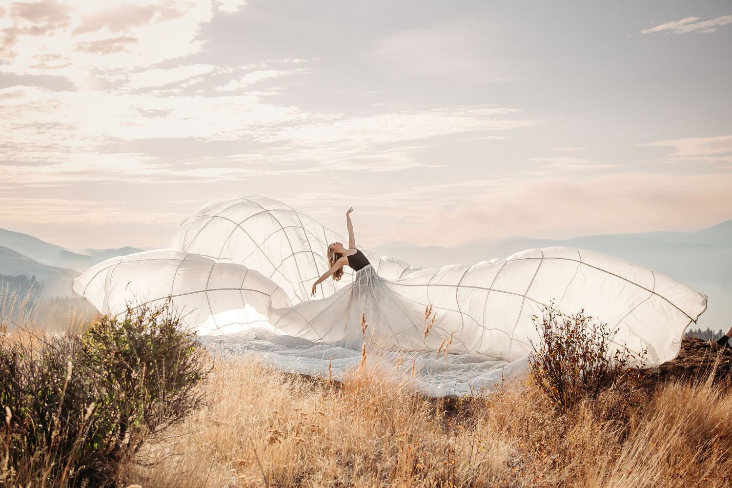 Figure in flowing white dress stands dramatically in mountain field with billowing fabric creating ethereal wings at sunset.