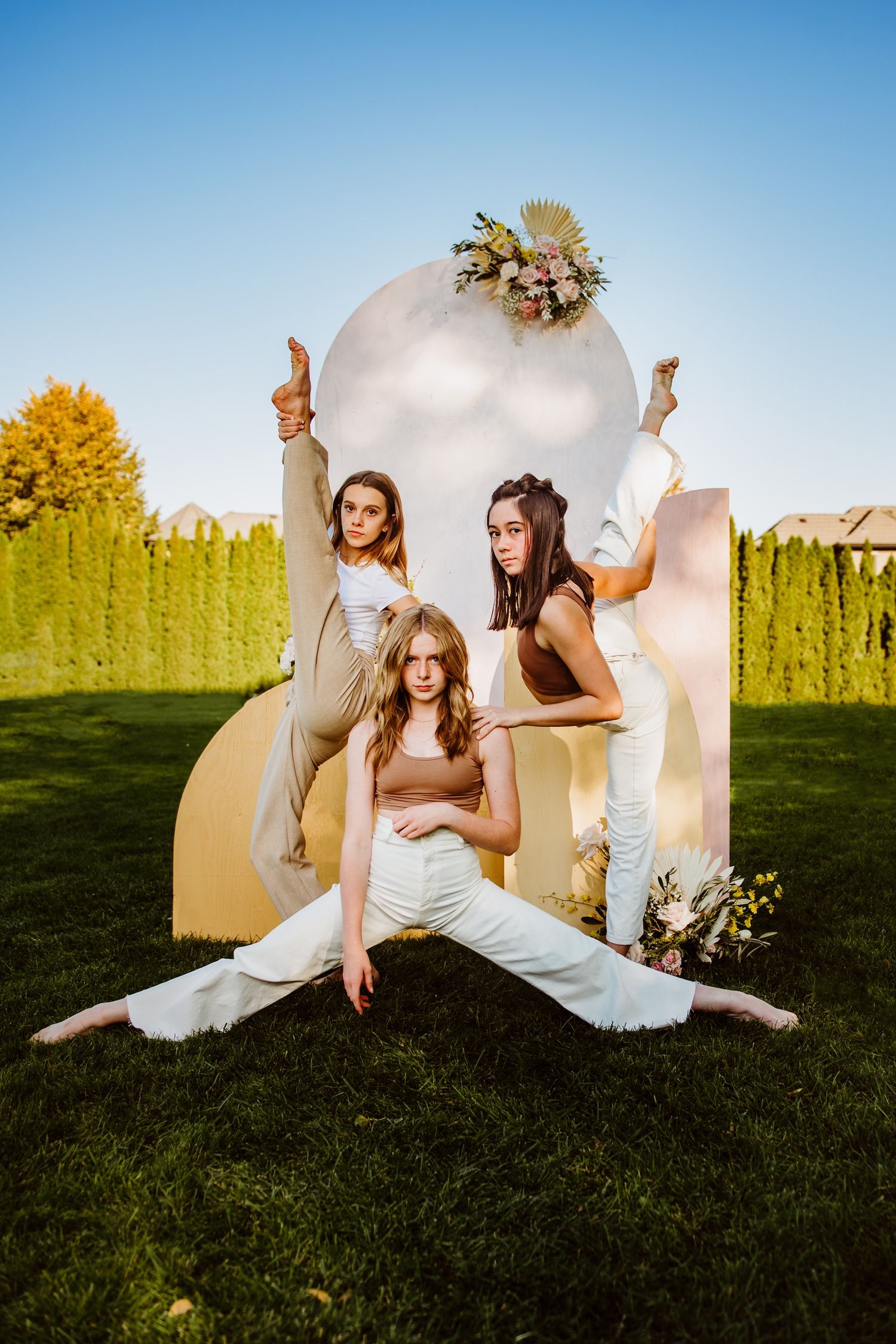 Three dancers in white outfits pose artistically against a decorative white arch backdrop in a sunny garden setting.