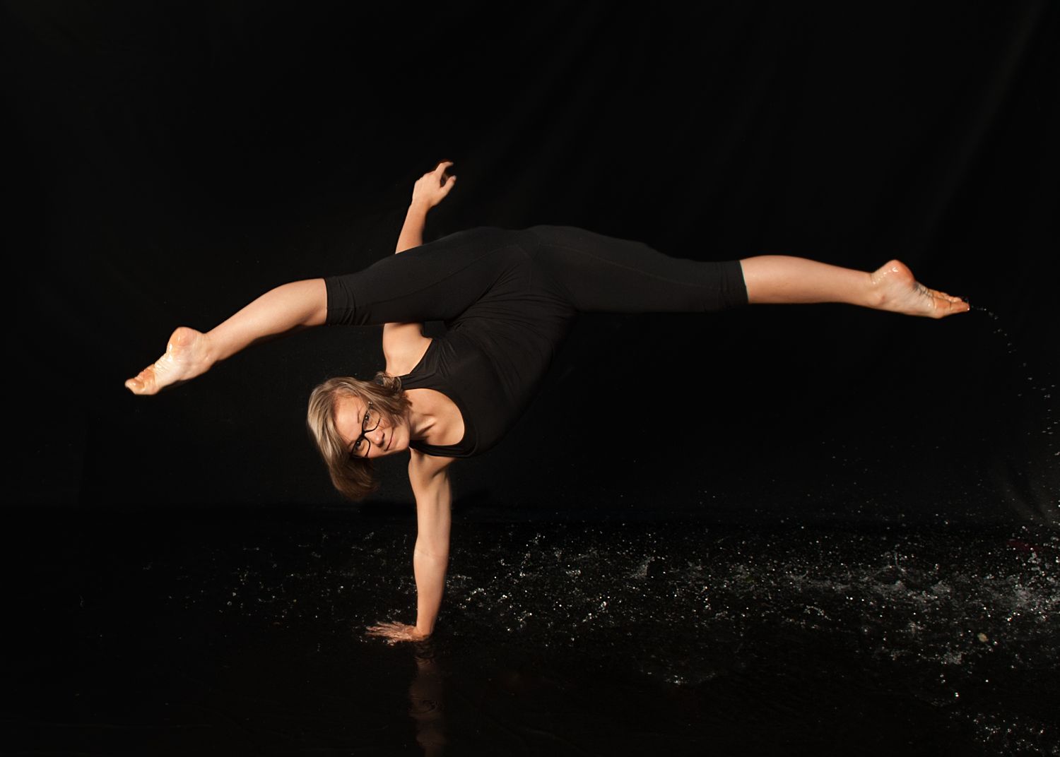 Dancer performs striking one-arm balance pose in black attire against dark background with dramatic lighting.