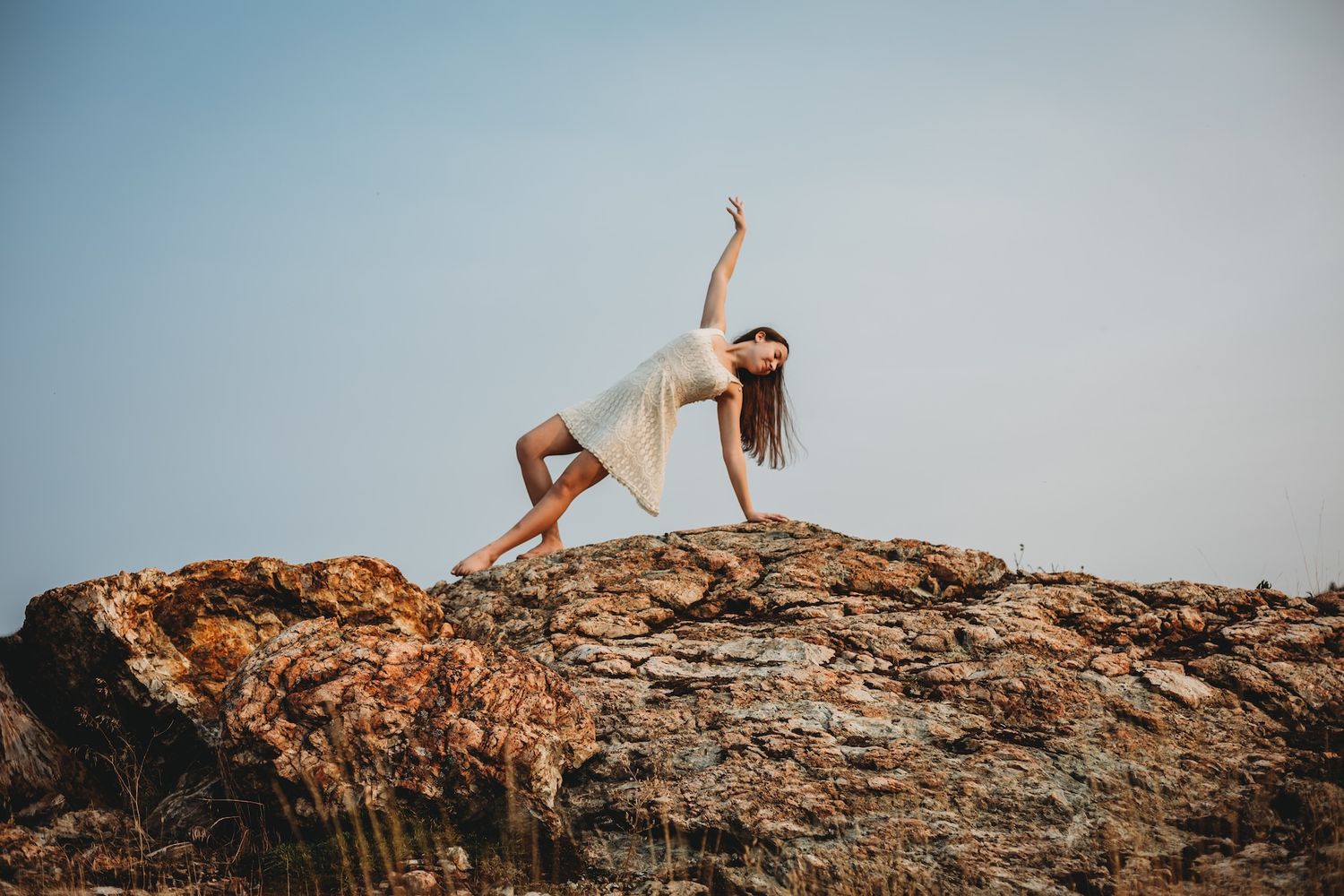 Dancer performs elegant pose on rugged rocky cliff against bright blue sky backdrop.