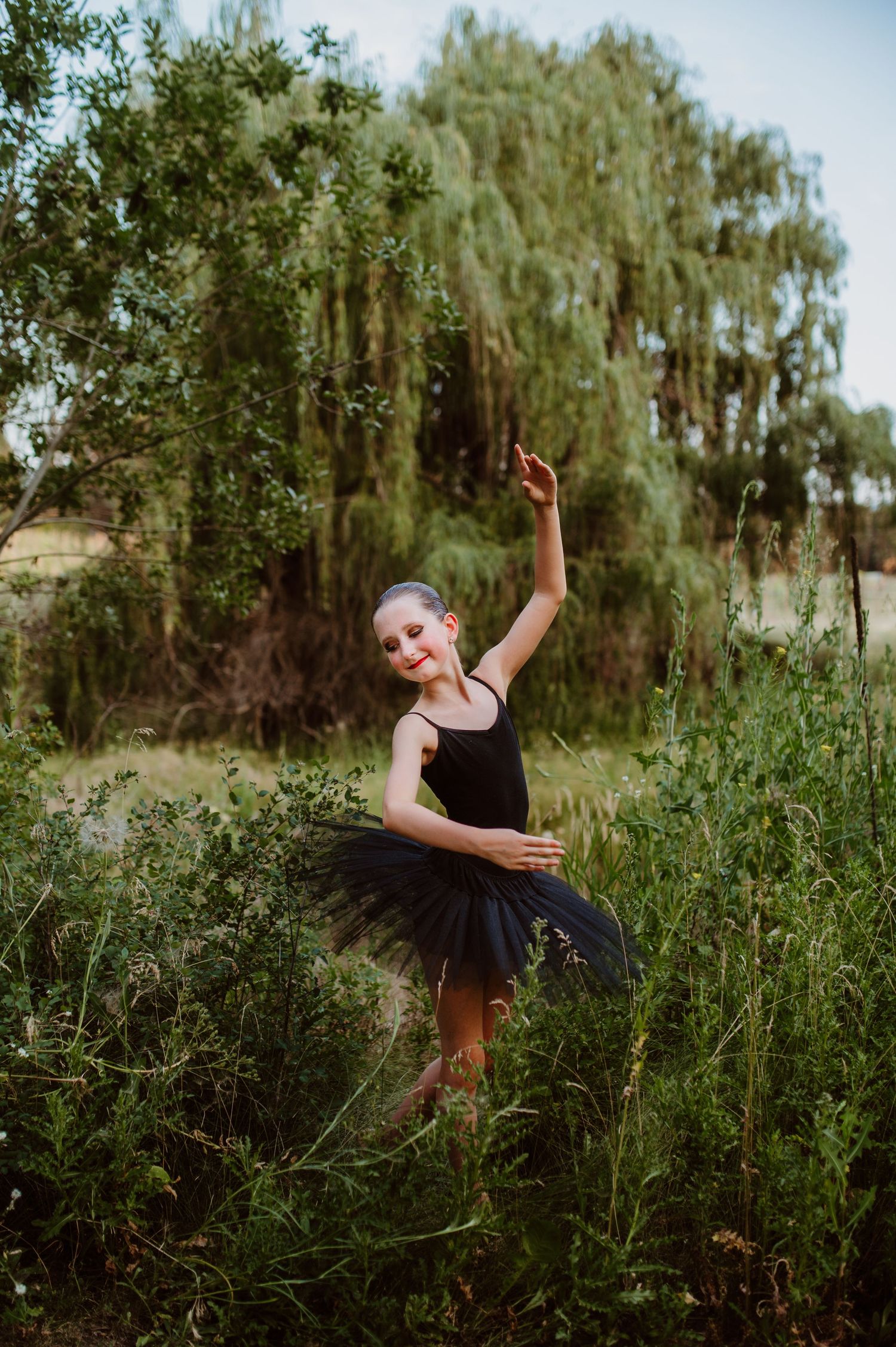 Young dancer in black tutu performs graceful ballet pose outdoors among willow trees and tall grass.