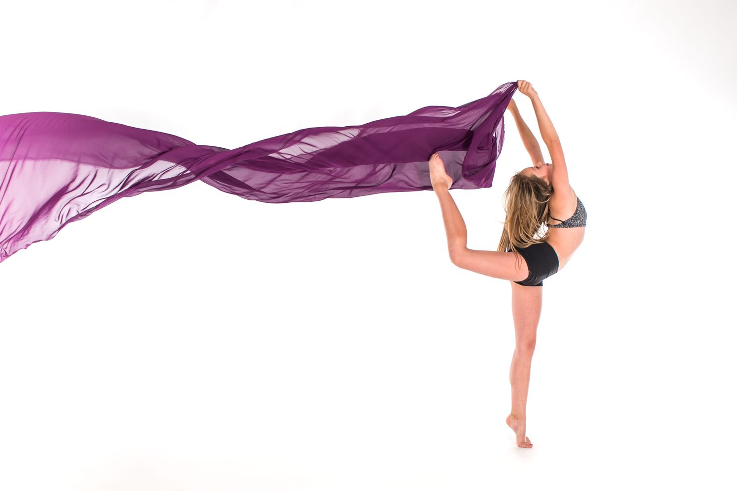 Dancer in black leotard gracefully performs leg extension while holding flowing purple fabric against white background.