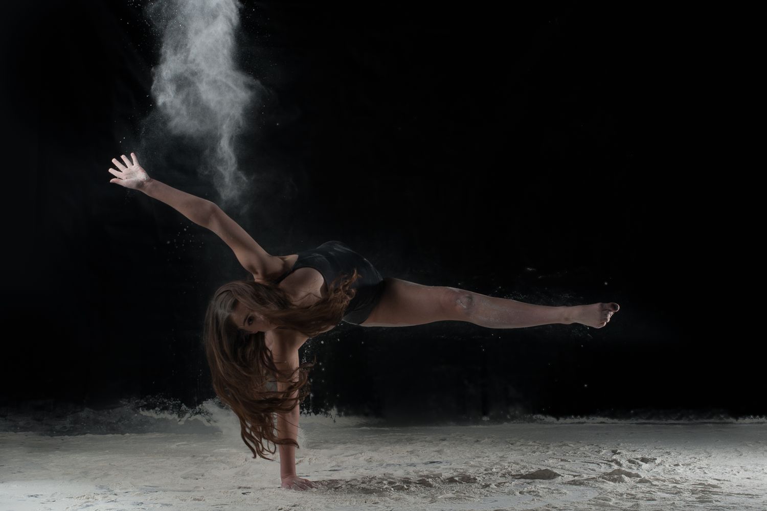 Contemporary dancer performs dynamic pose amid a cloud of white powder against a dark background.