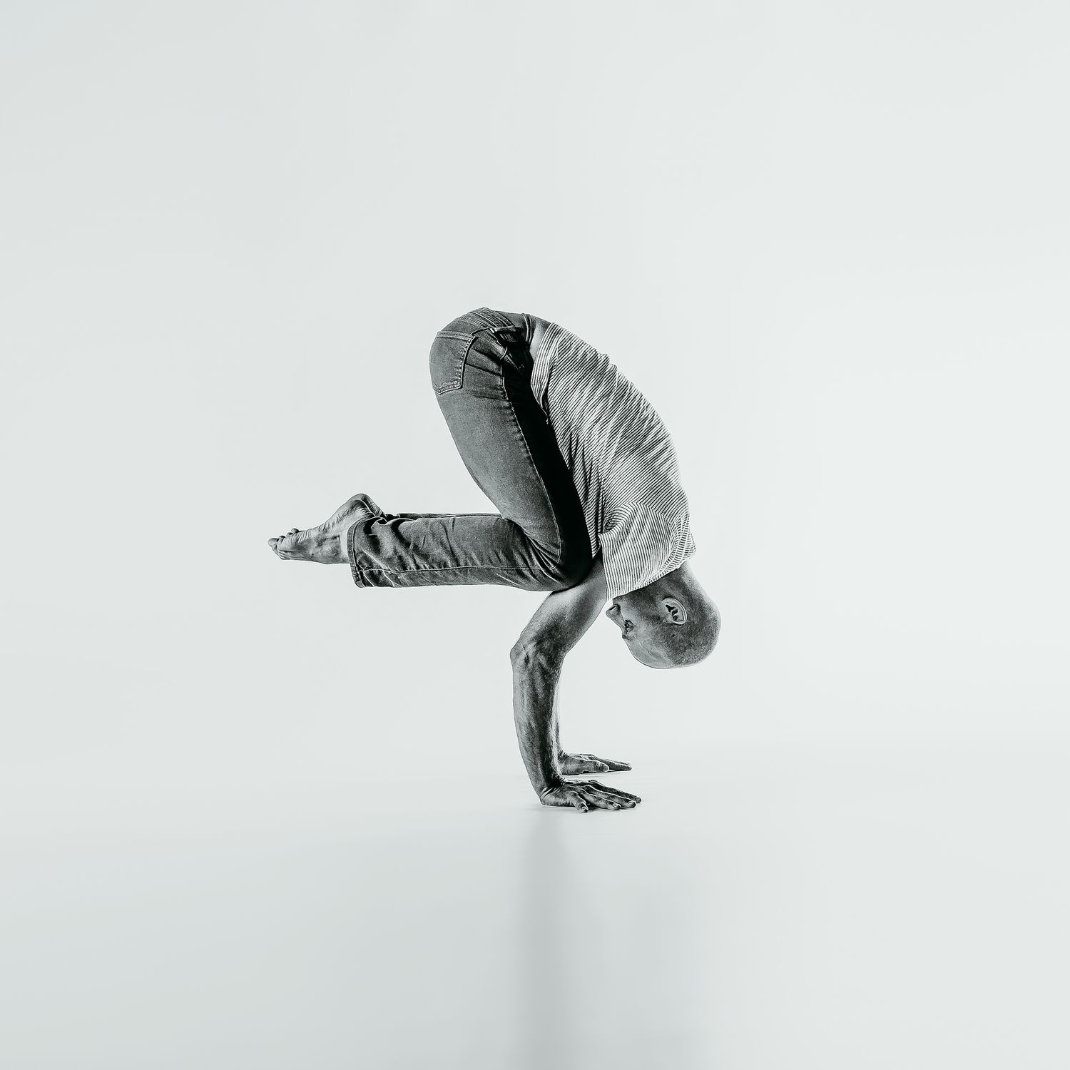 Black and white photo of figure performing a complex yoga crow pose balancing on hands against minimalist background.