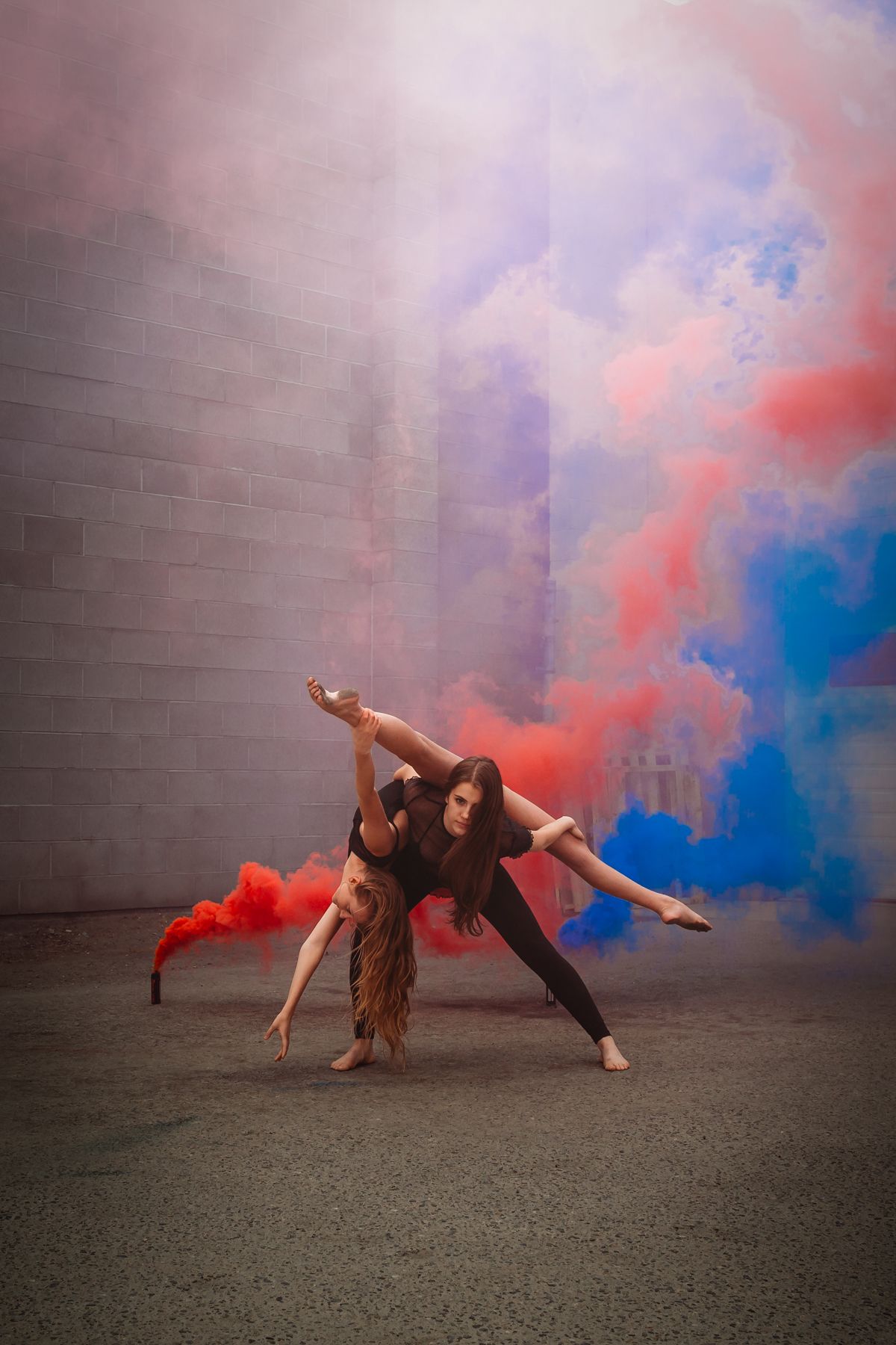 Contemporary dancers perform an expressive duet amid swirling red and blue smoke against an urban wall.