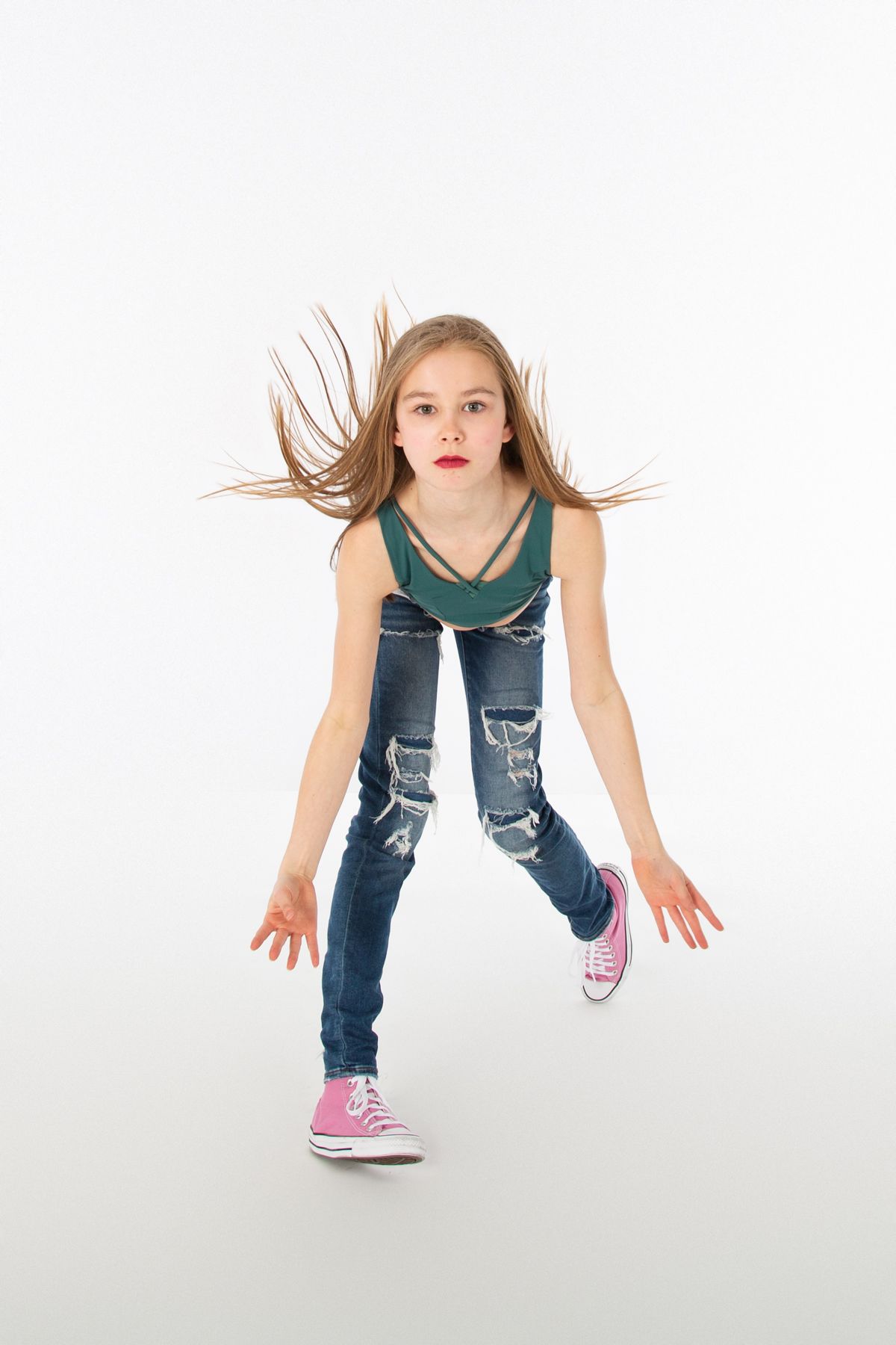 Dancer with flowing hair in distressed jeans and green top striking energetic pose on white backdrop.