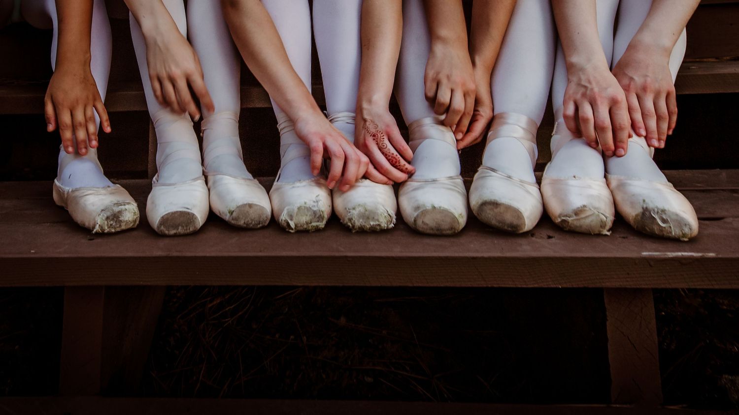 Ballet dancers sit together on wooden bench showing worn white pointe shoes and slippers after practice.