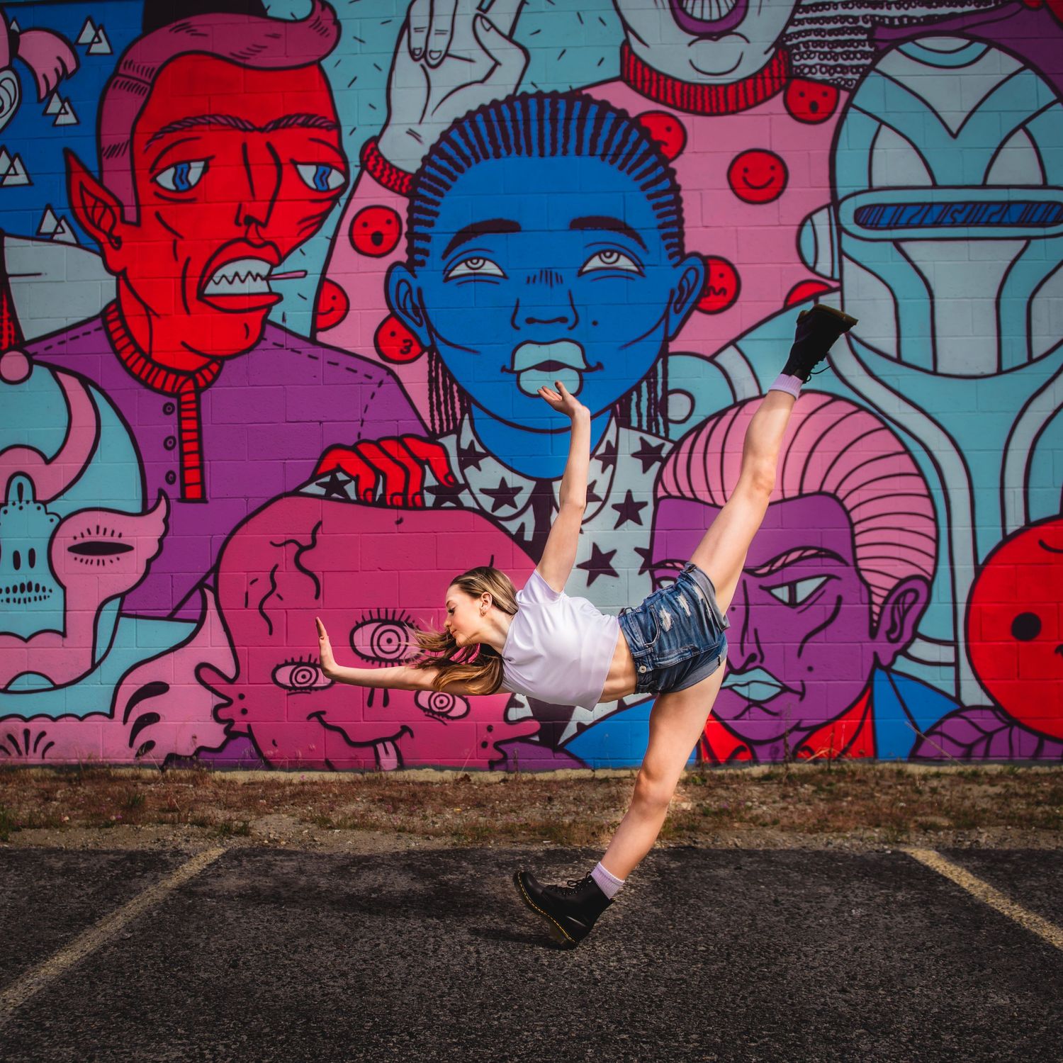 Dancer performs a dramatic kick move in front of colorful street art mural featuring vibrant cartoon faces.