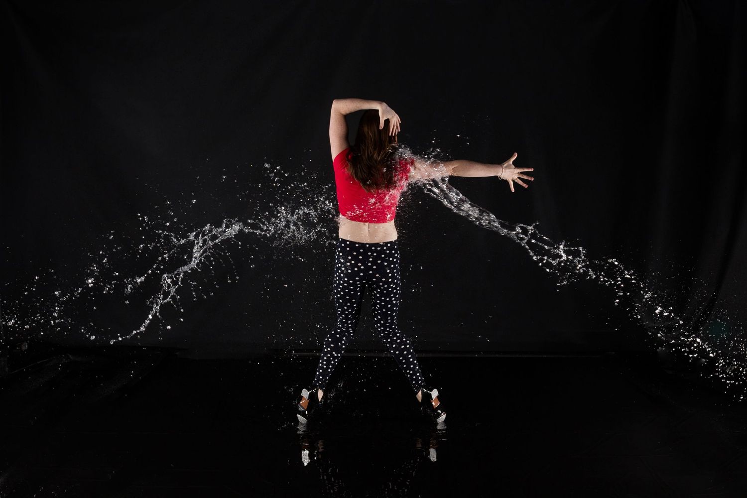 Dynamic dancer in red top and patterned pants creates dramatic water splash effect against black background.