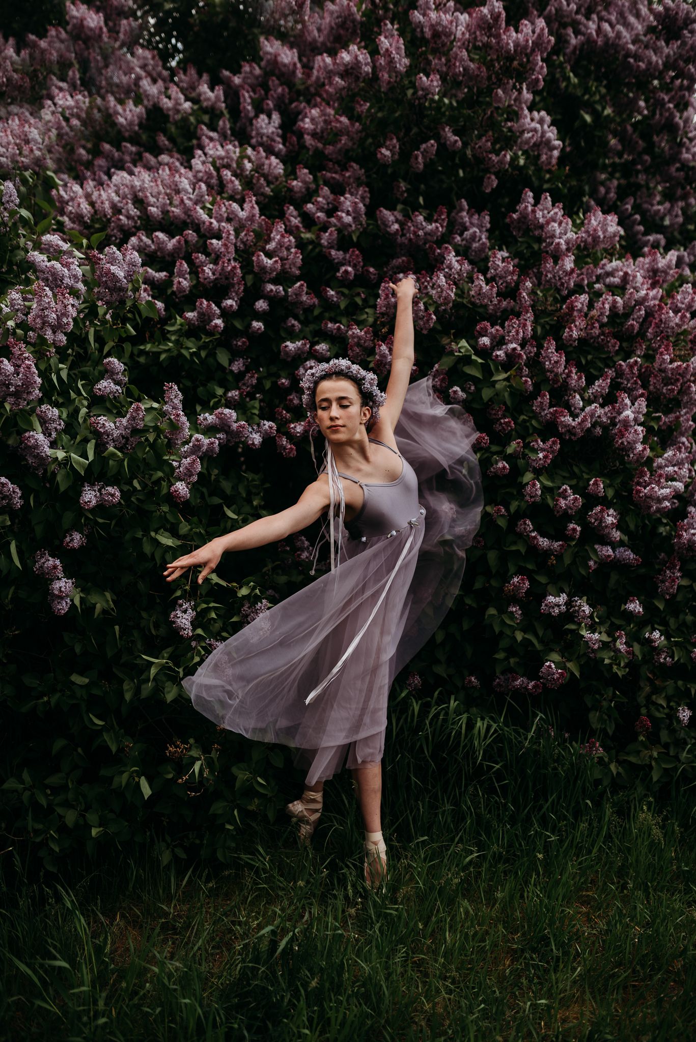 A ballet dancer in a flowing lavender dress performs an elegant pose against a backdrop of blooming lilac bushes.