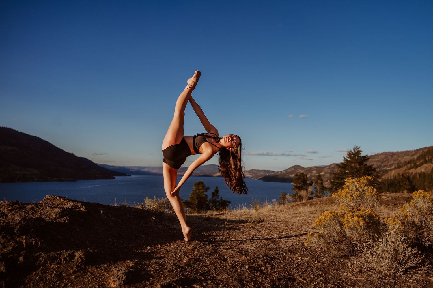A dancer performs a graceful pose against a scenic lakeside backdrop with mountains and blue sky at sunset.