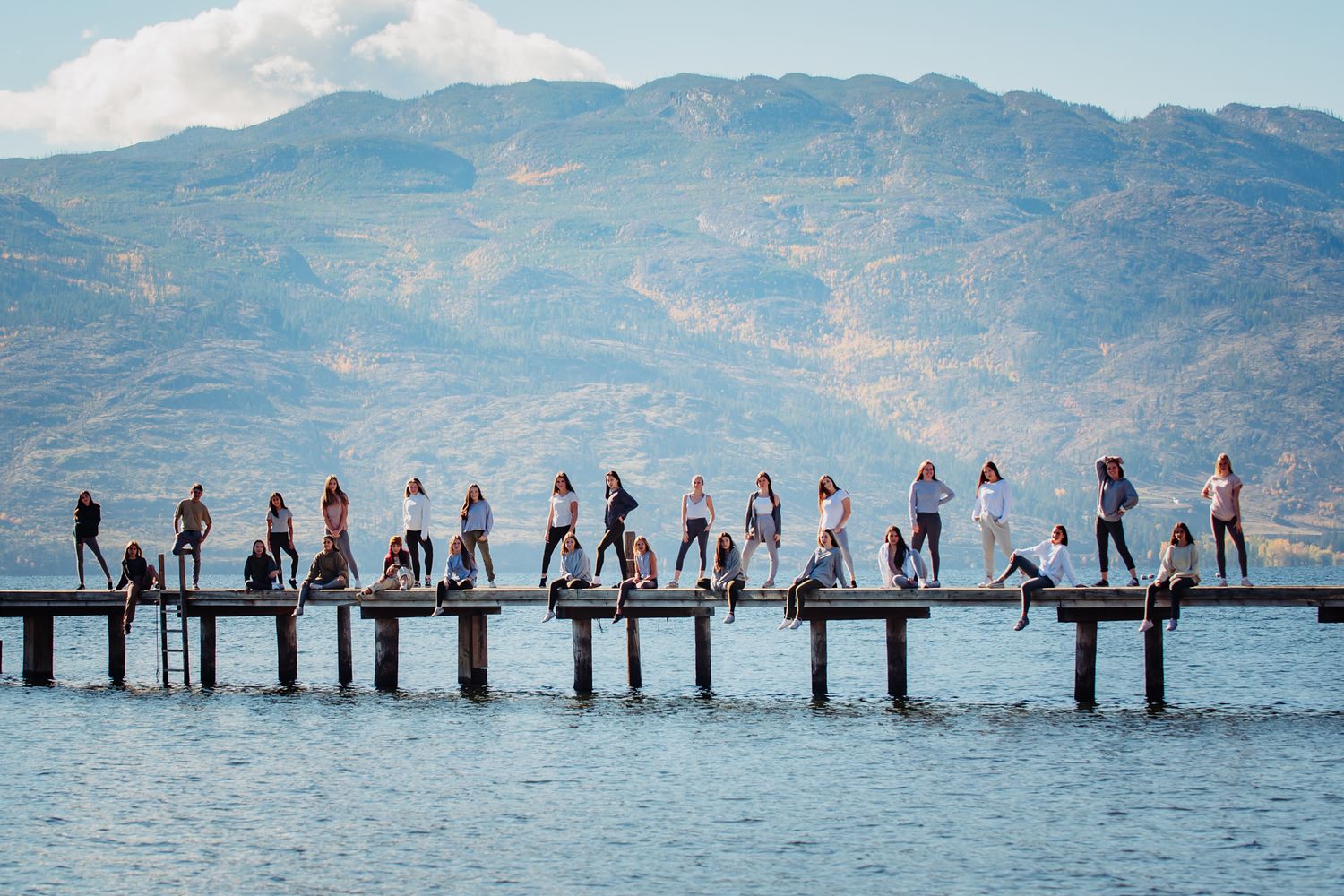 A group of people stand in silhouette on a long wooden pier over a lake with mountains in the background.