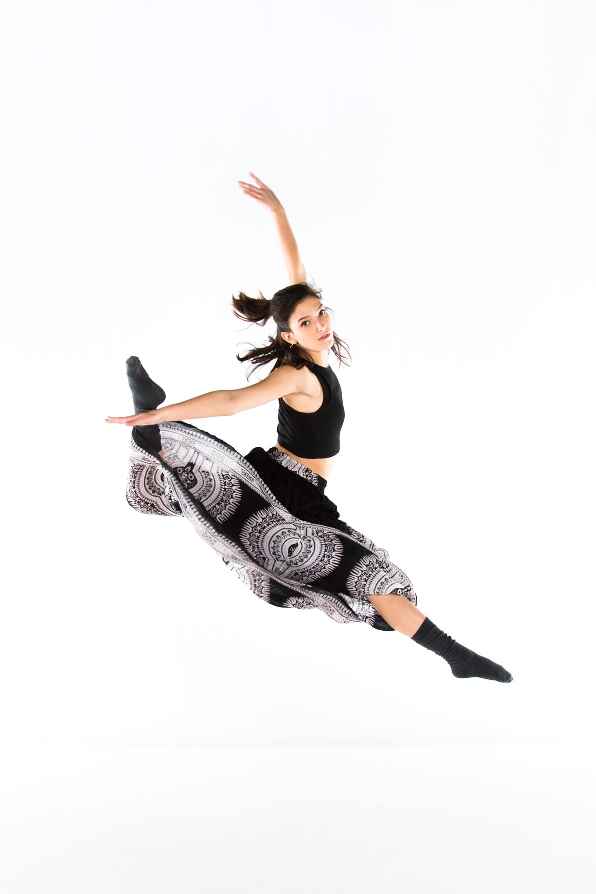 A dancer in patterned pants performs a dynamic split jump pose against a white studio backdrop.