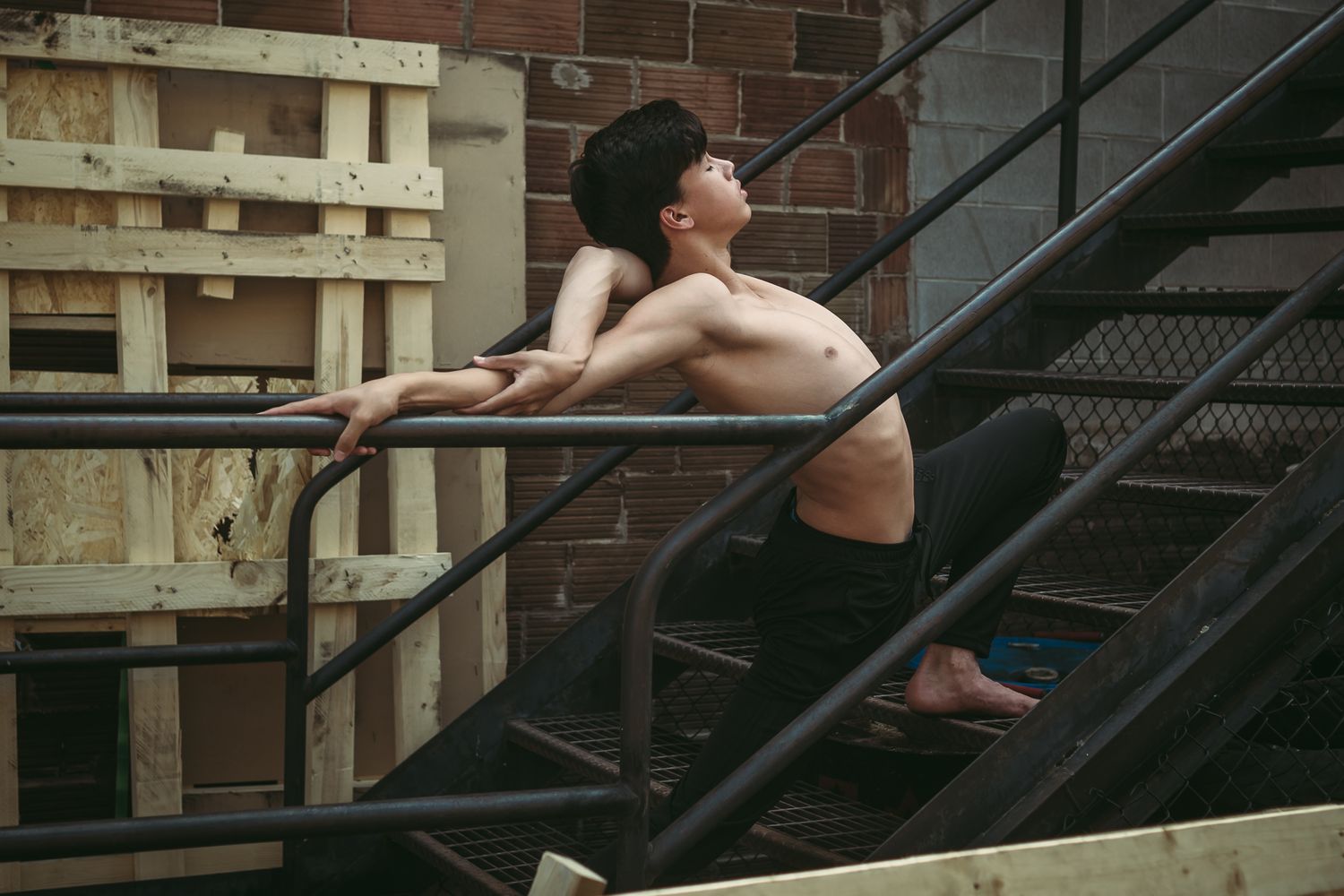 Shirtless dancer strikes elegant pose on metal staircase against brick wall in atmospheric urban setting.