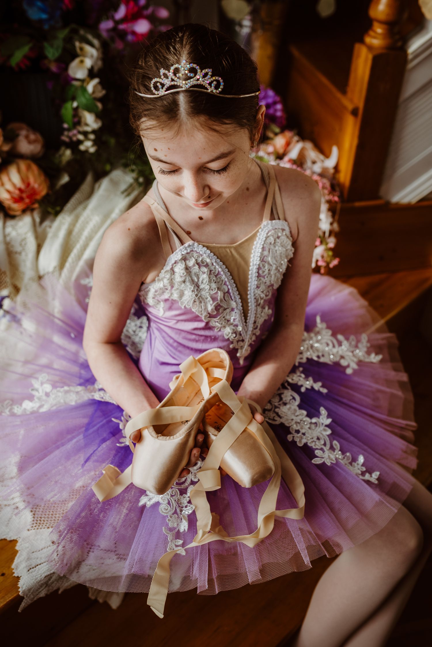 A young dancer in a purple tutu dress and tiara sits holding ballet pointe shoes surrounded by floral decorations.