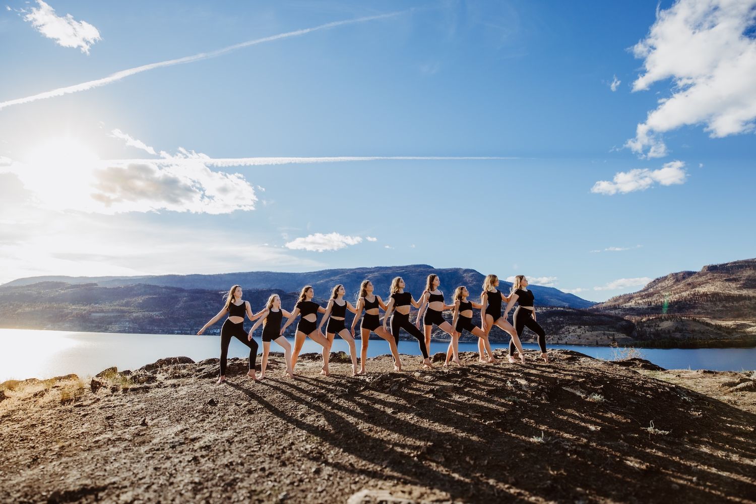 Group performs synchronized yoga poses on cliff edge overlooking scenic mountain lake at sunset.