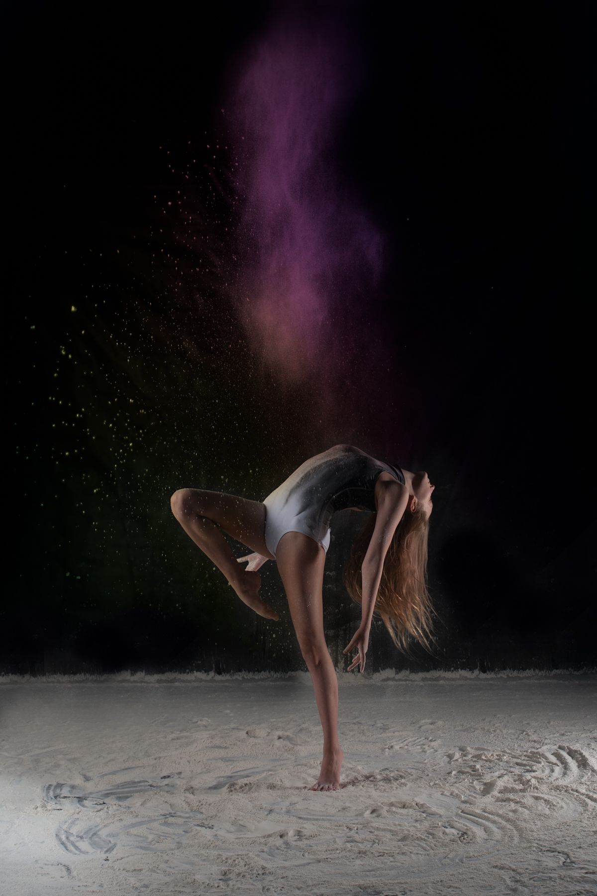 A dancer performs a dramatic backbend pose with purple smoke effects against a dark backdrop with white powder on floor.
