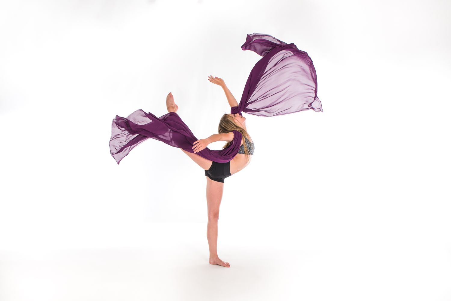 Dancer in black leotard performs graceful high kick with flowing purple fabric creating dramatic movement effect.
