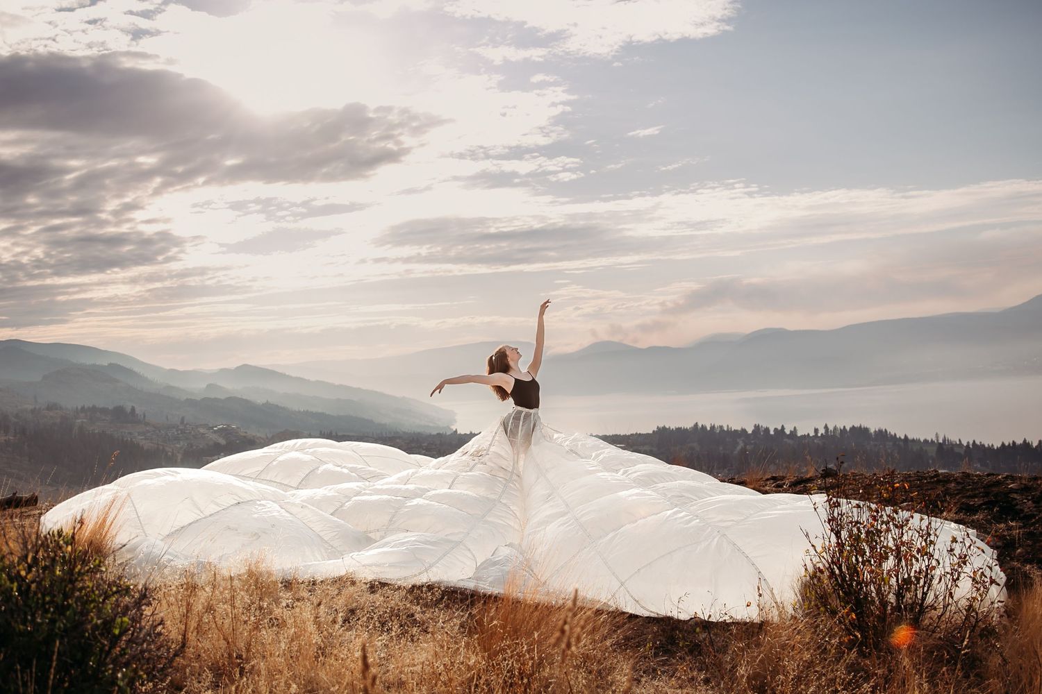 A bride in a flowing white dress stands triumphantly on a mountain overlook during sunset with dramatic clouds above.