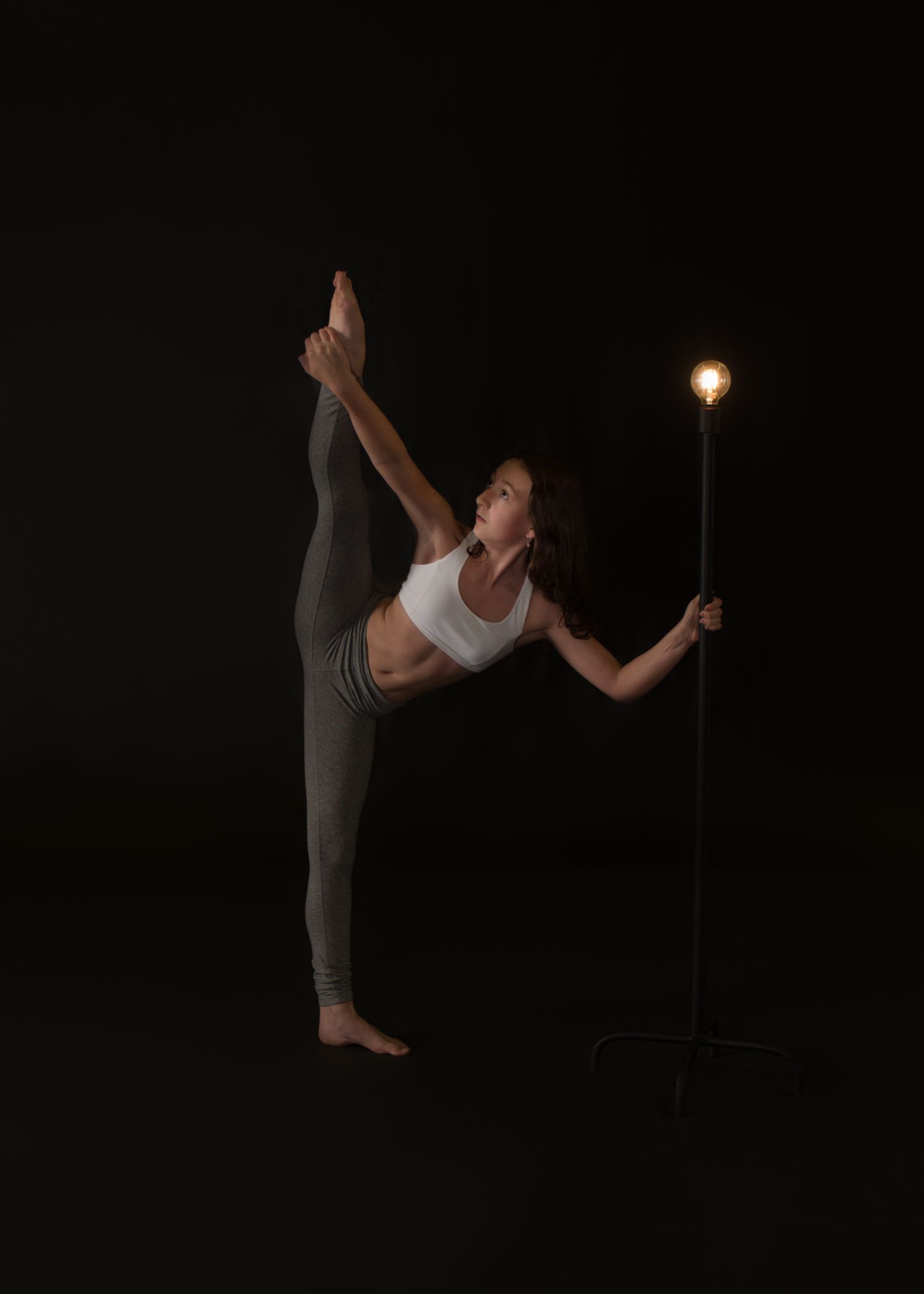 Dancer performs standing split pose while holding illuminated light stand against dark background in dramatic artistic photo.