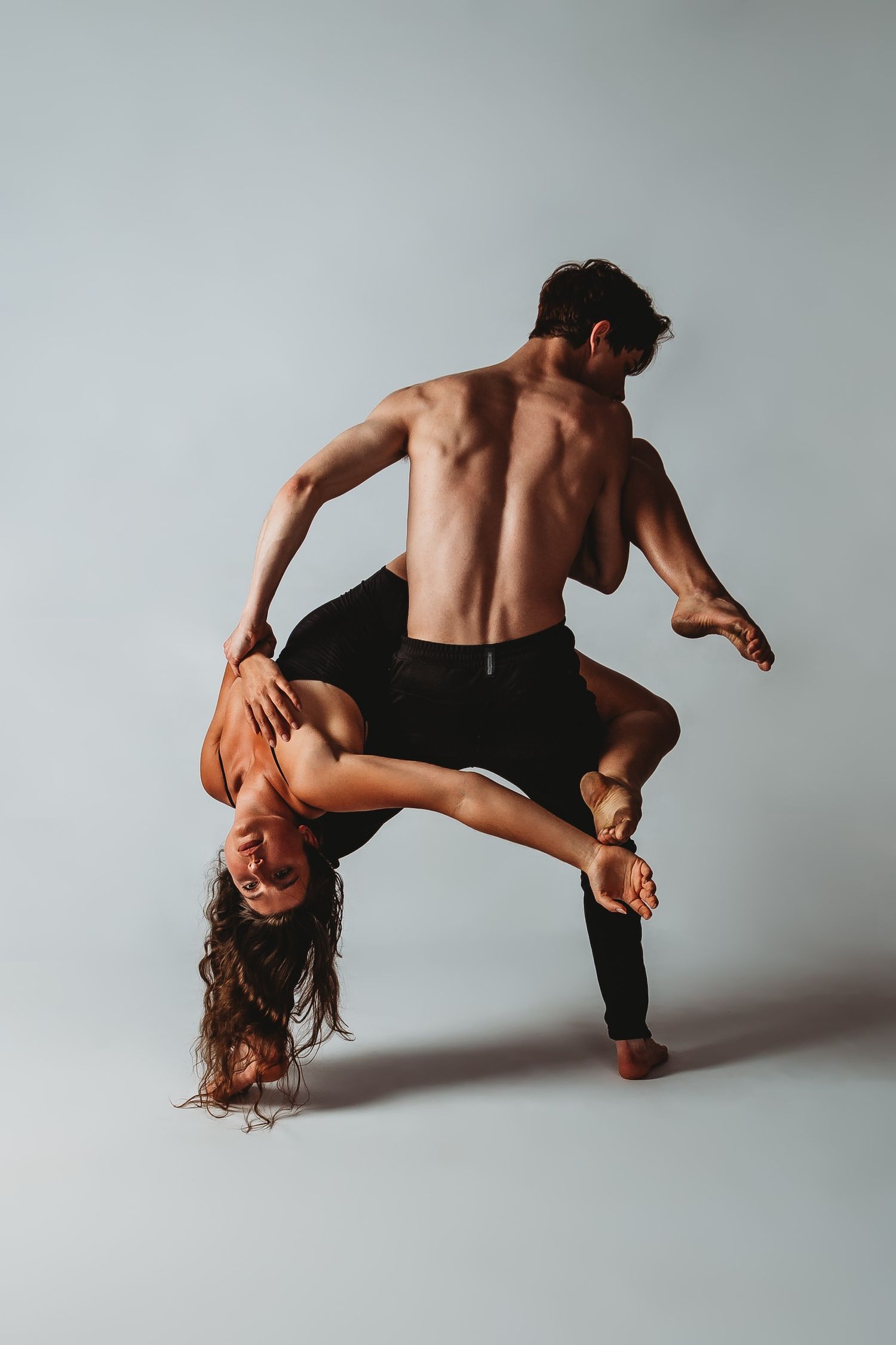 Two dancers perform an intimate contemporary dance pose against a minimalist gray backdrop.