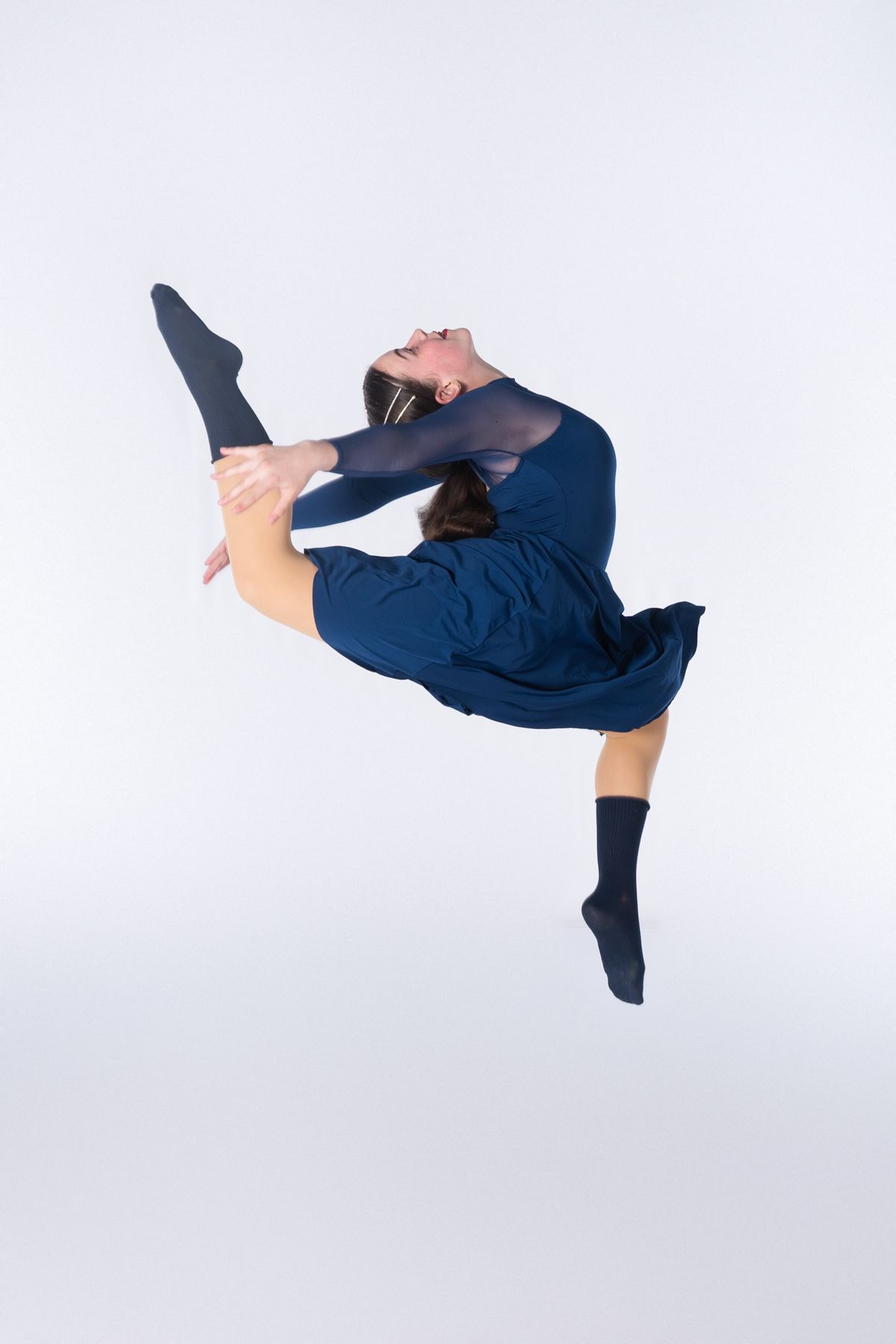 Modern dancer in navy blue dress leaps gracefully through the air against white background.