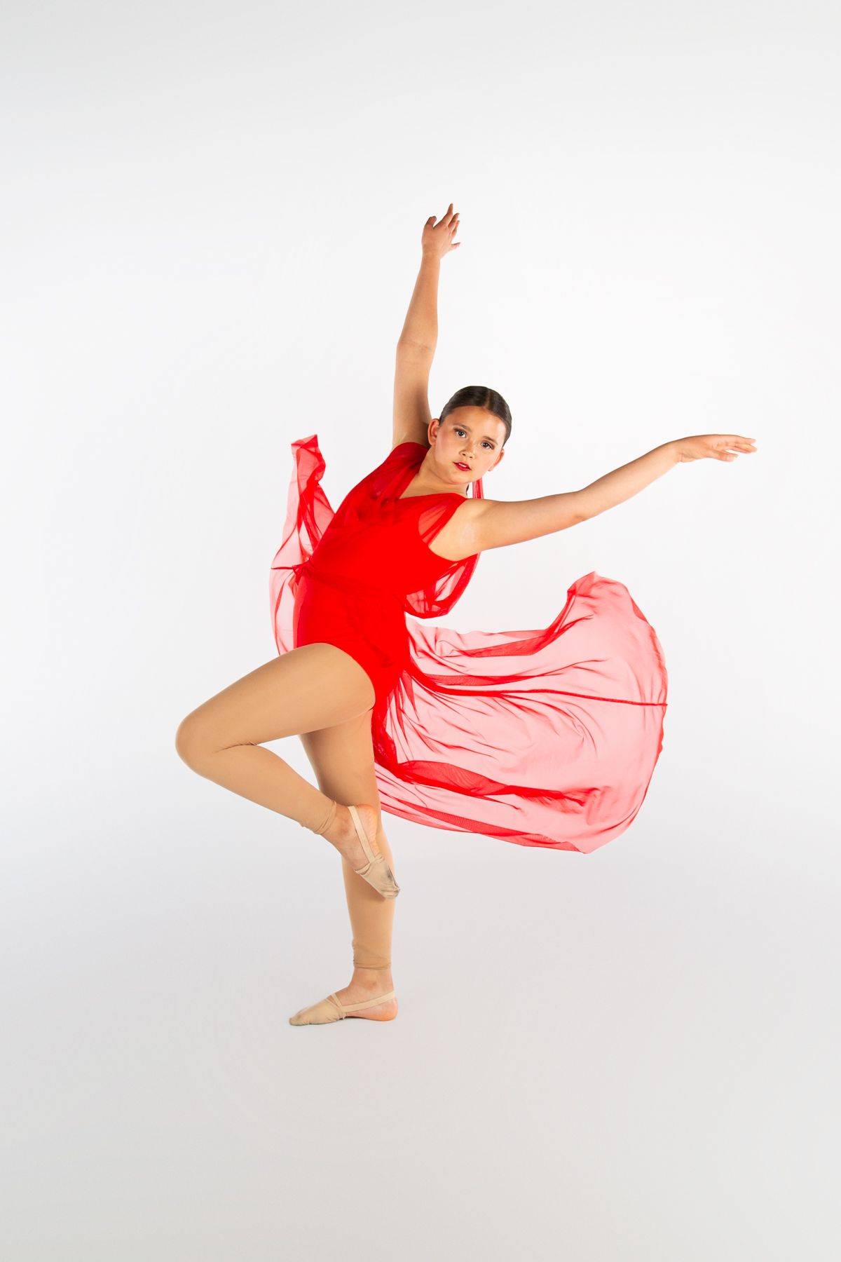 A dancer in a flowing red costume performs an elegant pose with one leg raised and arms extended against a white backdrop.