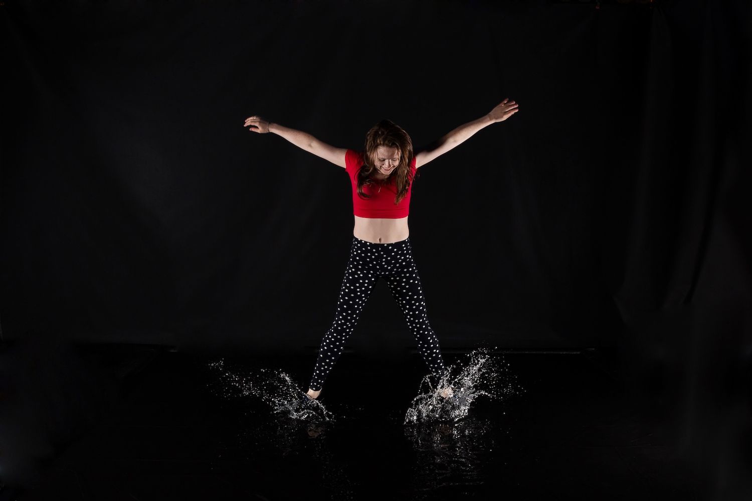 Athletic dancer in red top splashes water while jumping with outstretched arms on dark backdrop.