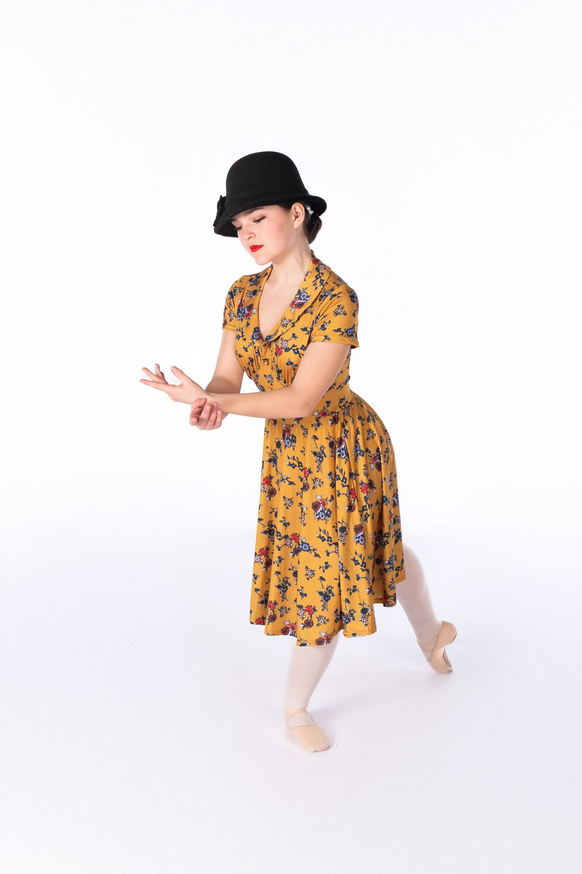 Dancer in vintage yellow floral dress and black bowler hat strikes theatrical pose in ballet shoes.