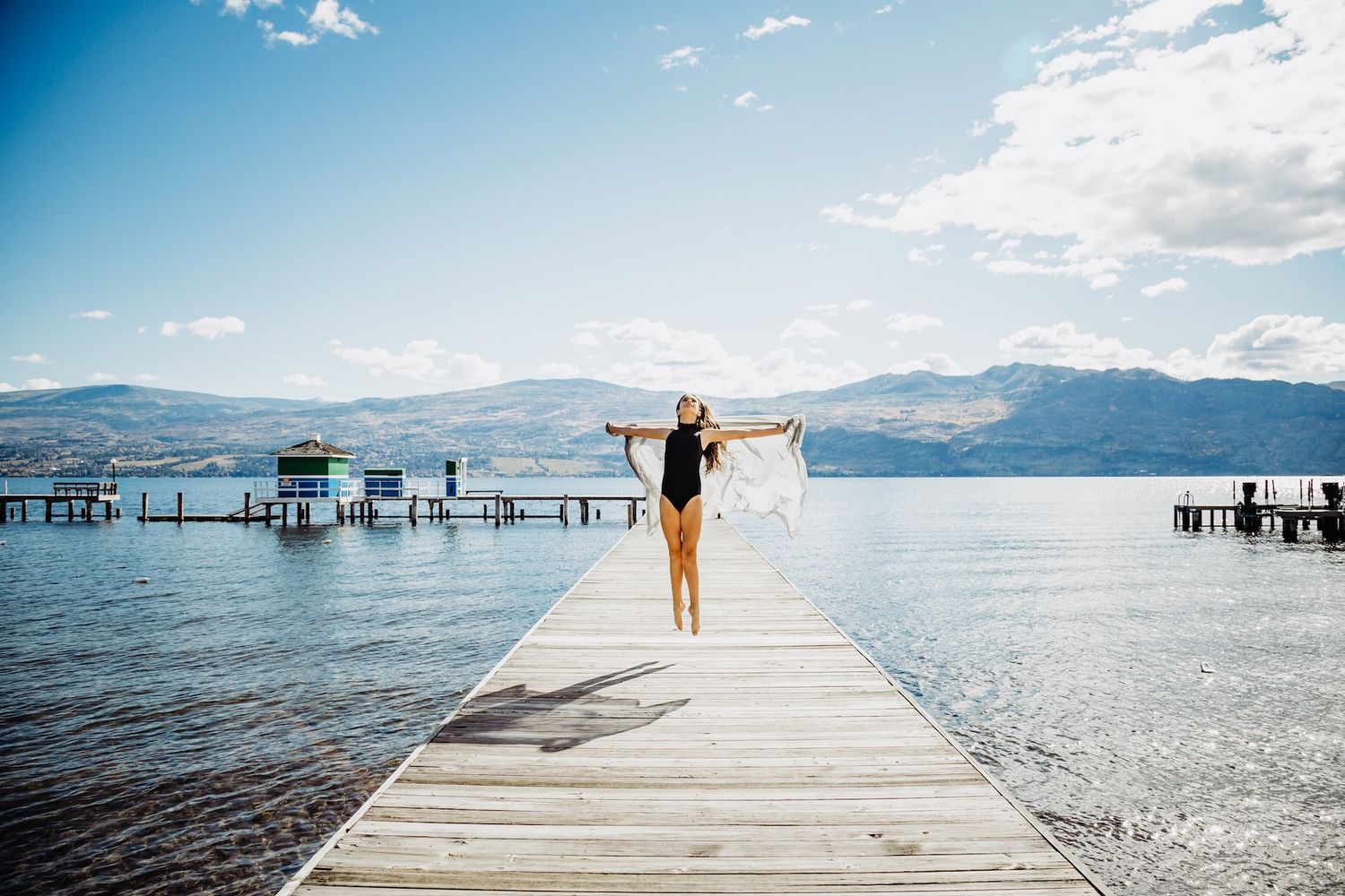 A wooden pier stretches into a tranquil lake surrounded by mountains on a sunny day with scattered clouds.