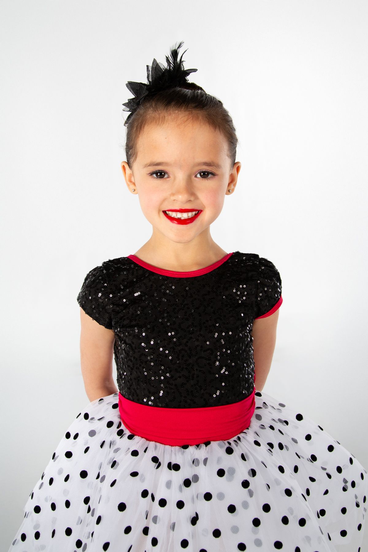 A performer in a sparkly black and red dance costume with a white polka dot tulle skirt stands against a white background.