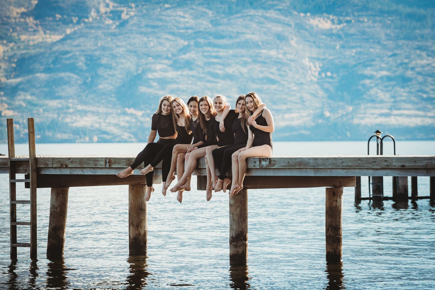 A group of friends sit together on a wooden dock over tranquil blue water on a sunny day.