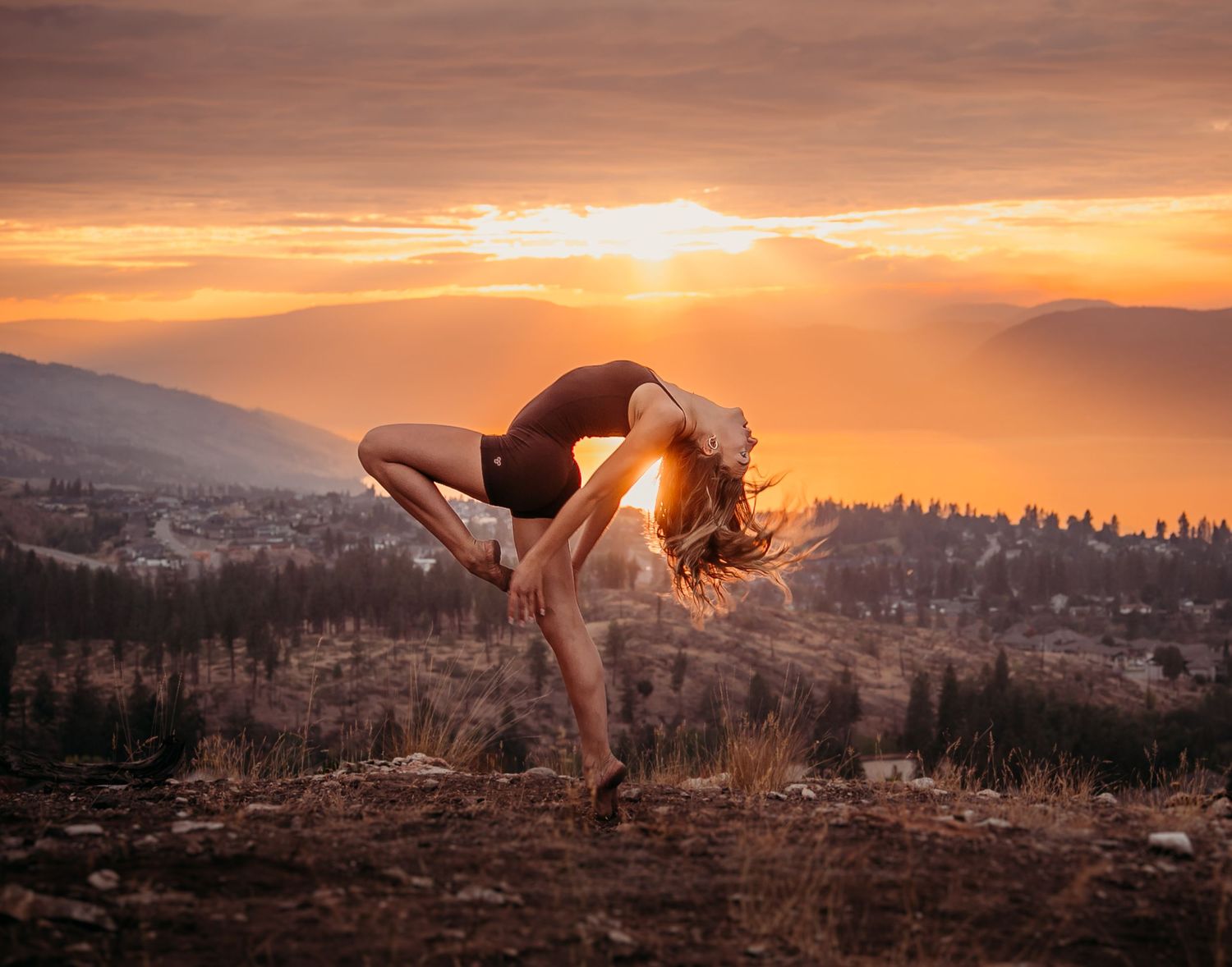 A graceful dancer performs a backbend pose against a stunning mountain sunset backdrop in vibrant orange hues.