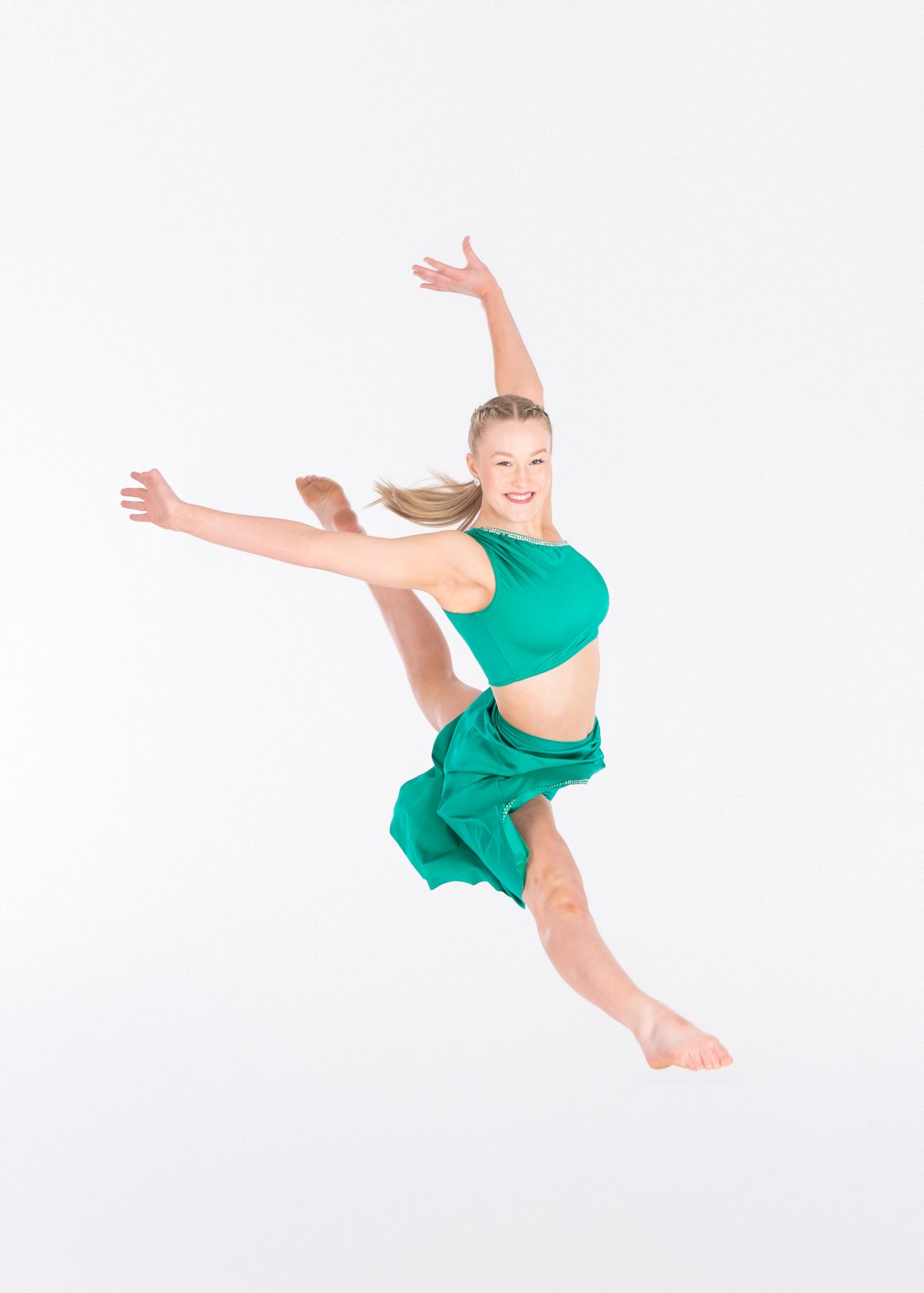 Dancer in emerald green outfit performs a graceful leap with outstretched arms against white background.