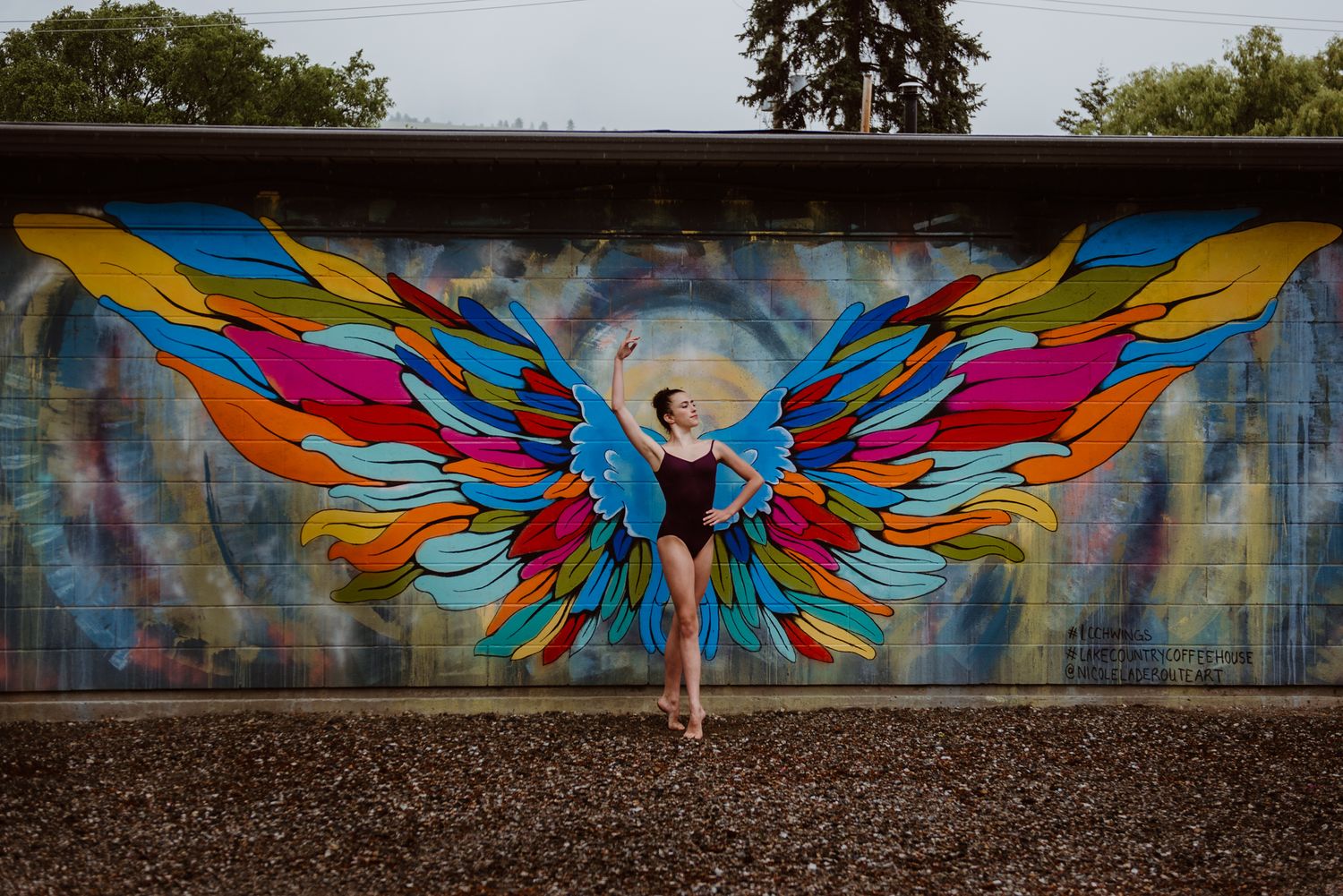 A dancer poses in front of a vibrant street art mural featuring colorful butterfly wings on a concrete wall.