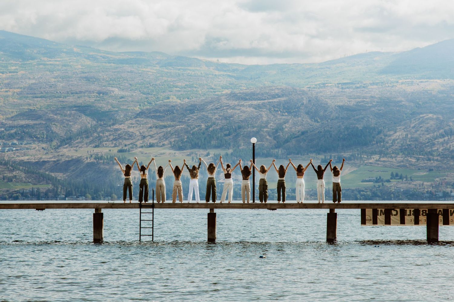 Silhouetted figures stand with raised arms on a wooden pier overlooking a scenic mountain lake on a cloudy day.