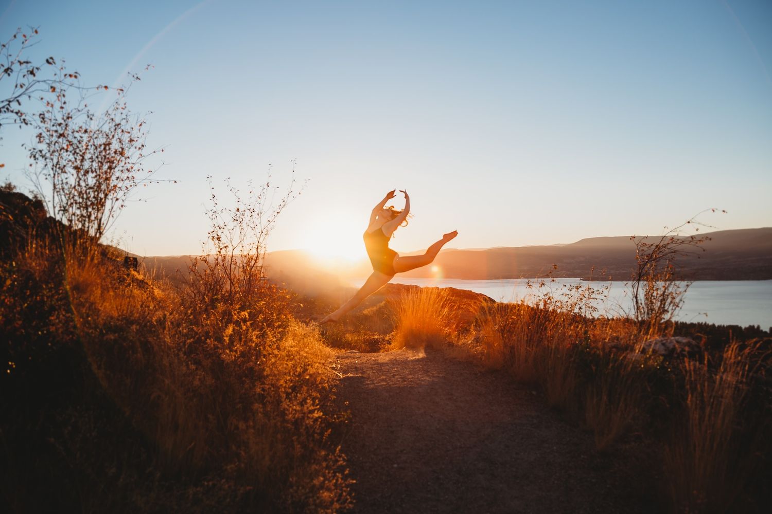 Silhouetted figure jumps joyfully at sunset along a scenic lakeside trail in warm golden light.