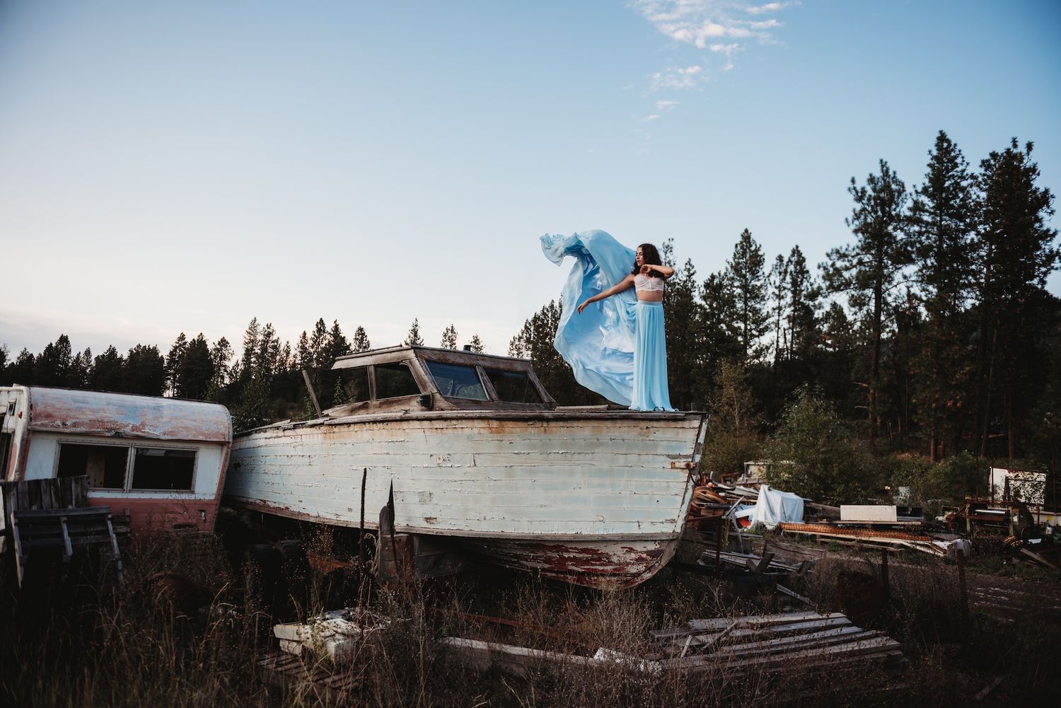 A snowboarder performs an aerial trick off an old abandoned boat in a wooded winter setting.